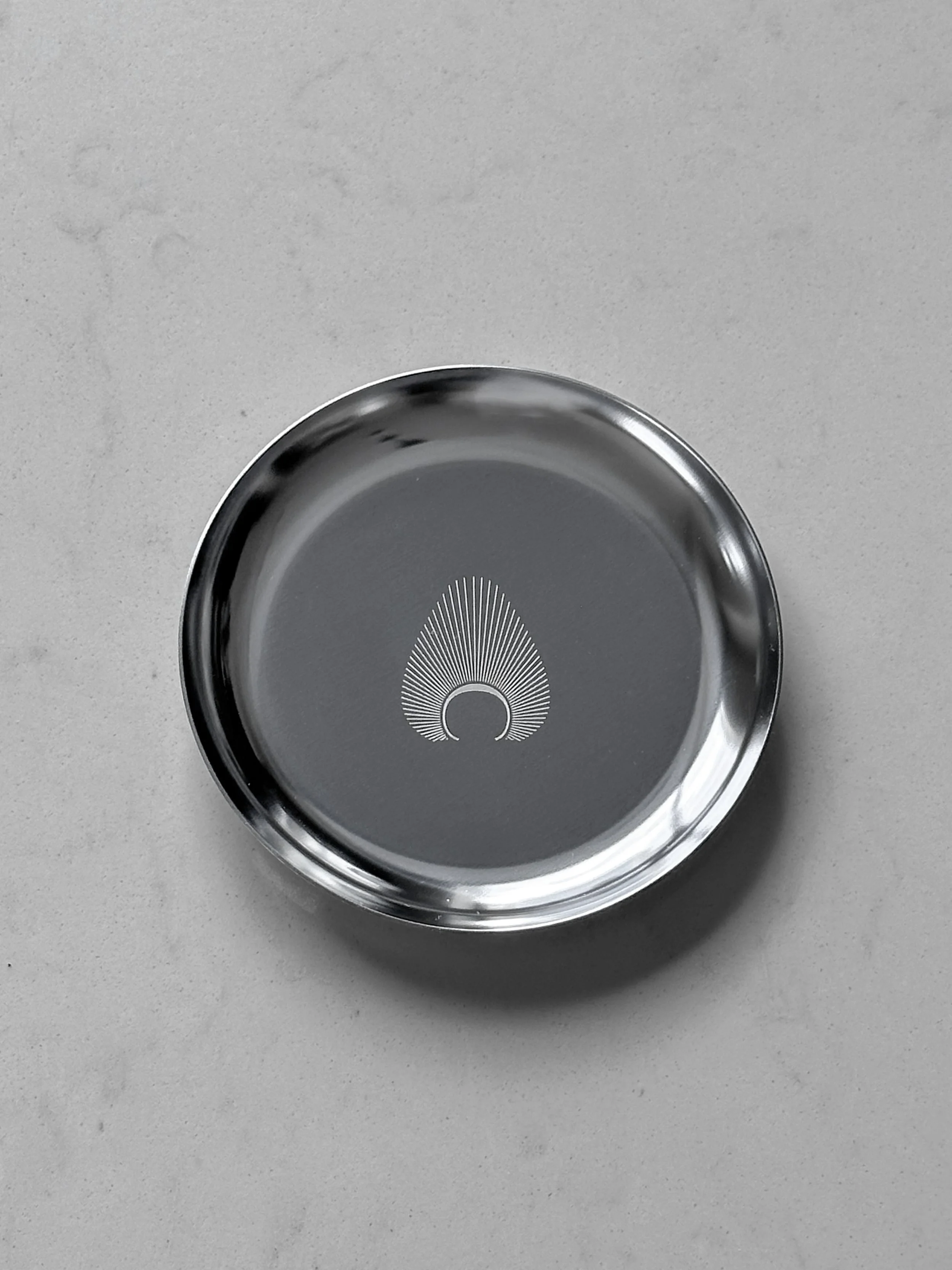 A round metal tray with a minimalist design of radiating lines from a small circle inside a larger circle on its center, placed on a light gray surface.