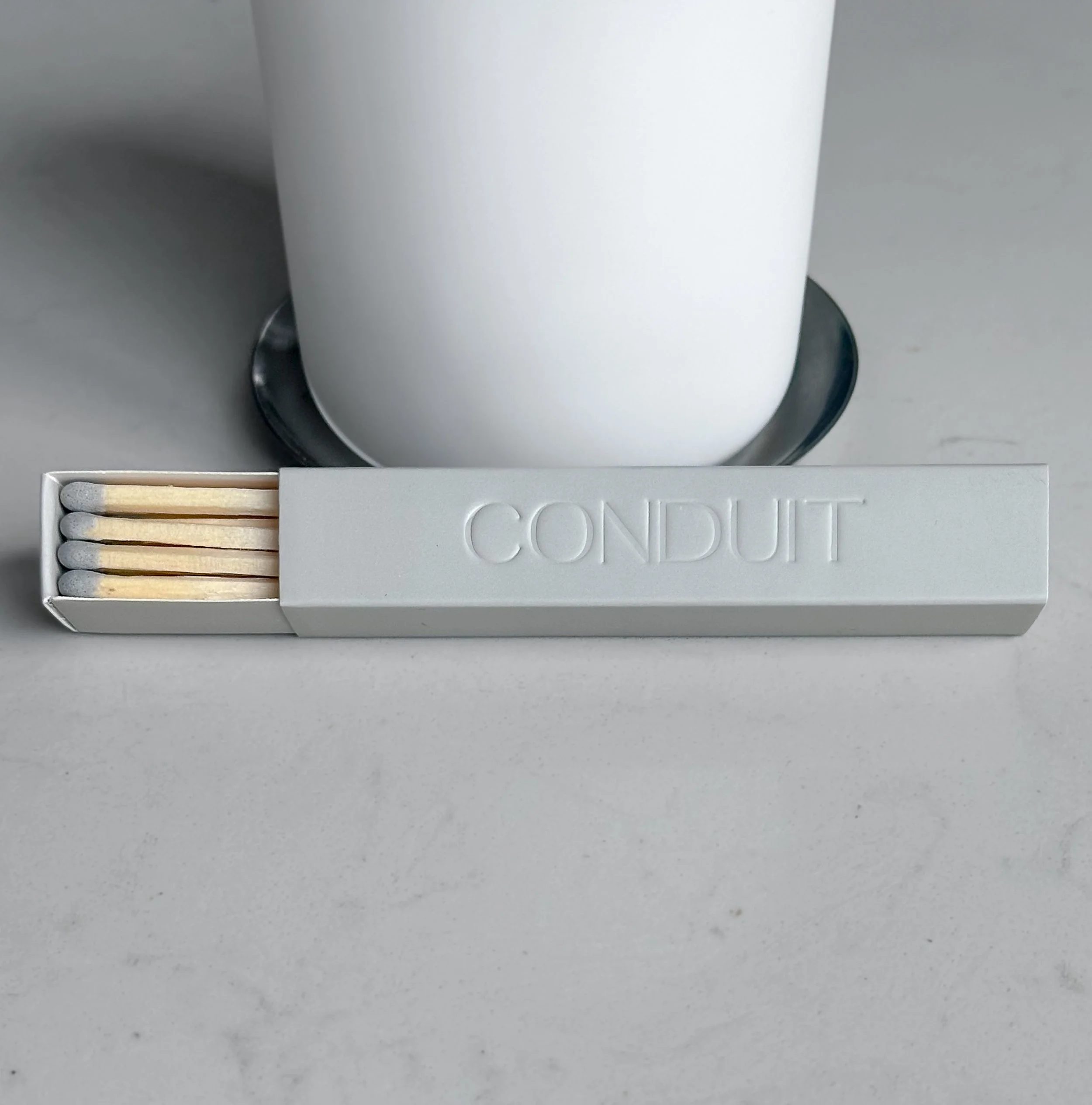 A white box of matches labeled 'CONDUIT' next to a white cup on a black plate, on a light gray surface.