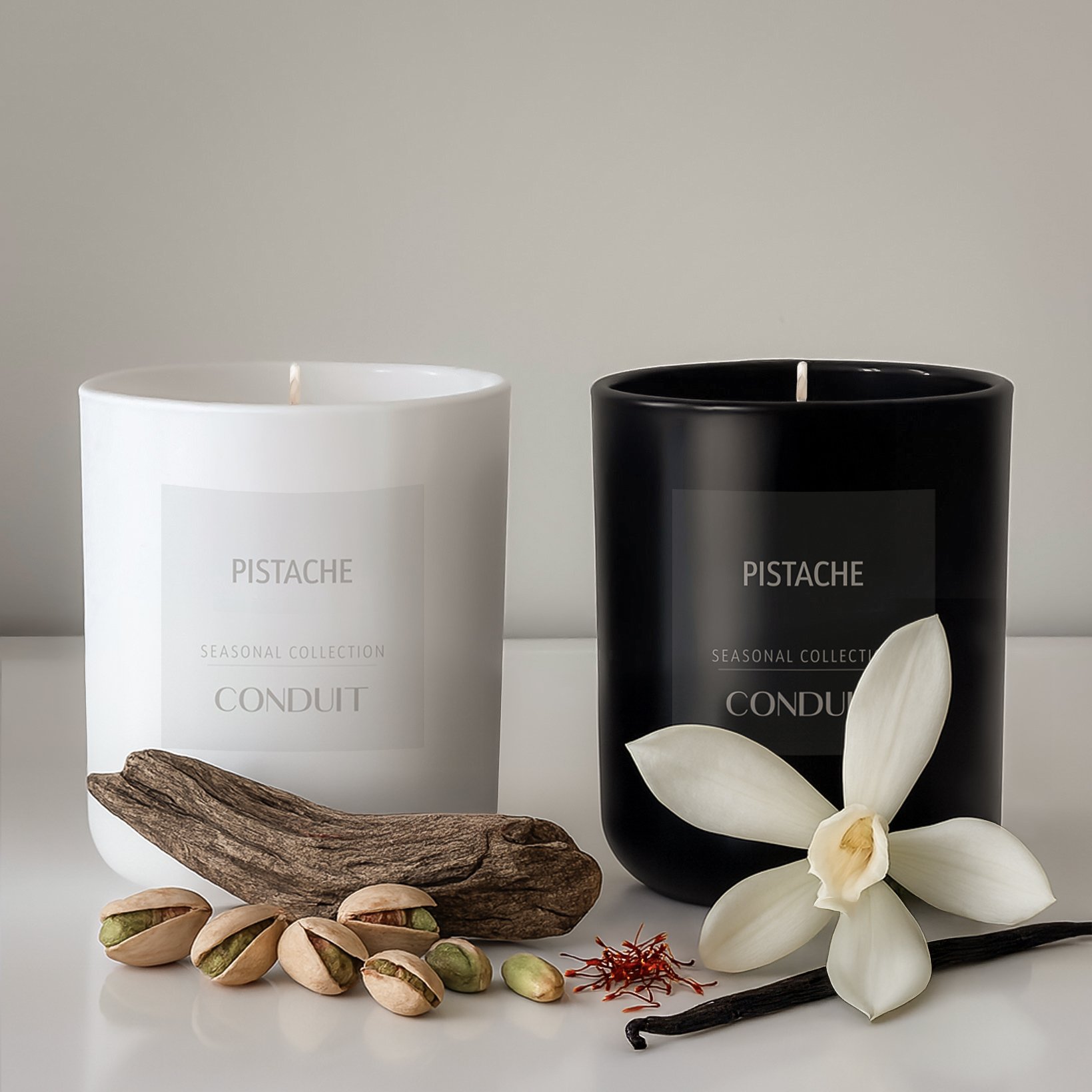 Two candles, one white and one black, labeled 'Pistache,' with a white pistache flower, a vanilla bean, pistachios, a piece of wood, and red saffron strands arranged in front.
