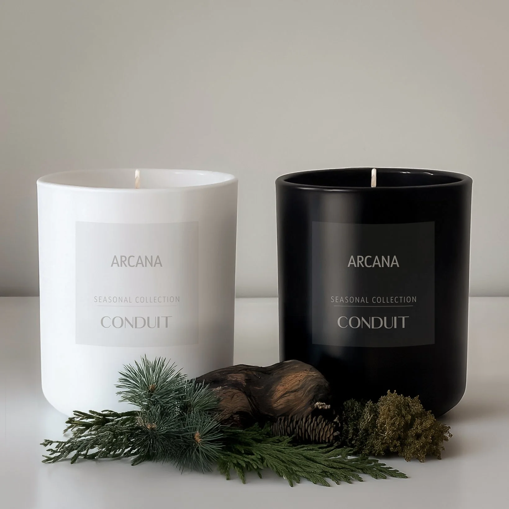 Two candles, one white and one black, labeled 'Arcana Seasonal Collection Conduit,' with decorative greenery and a piece of wood in front of them.