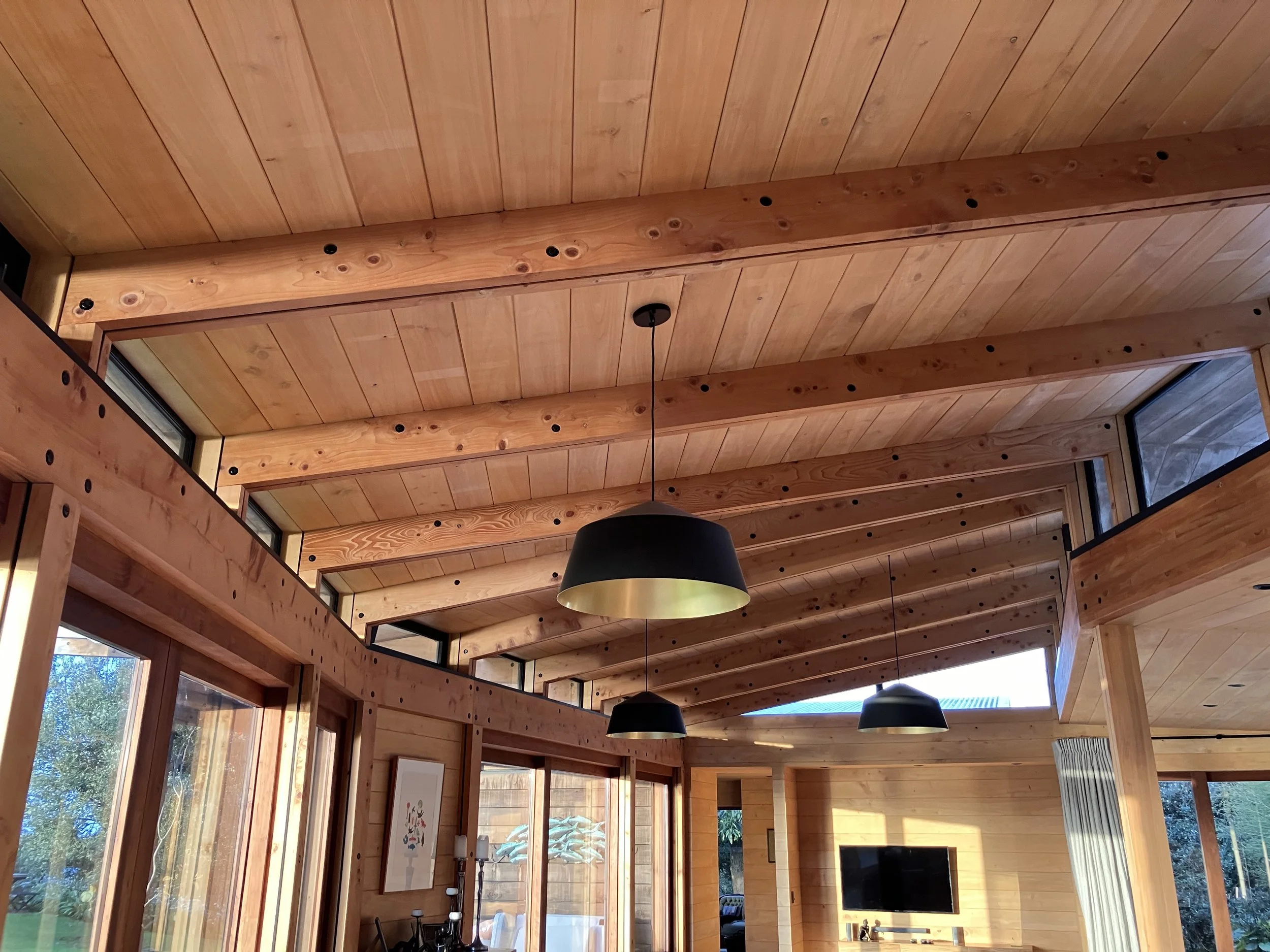 Wooden ceiling with large beams and hanging black pendant lights in a bright room with large windows.