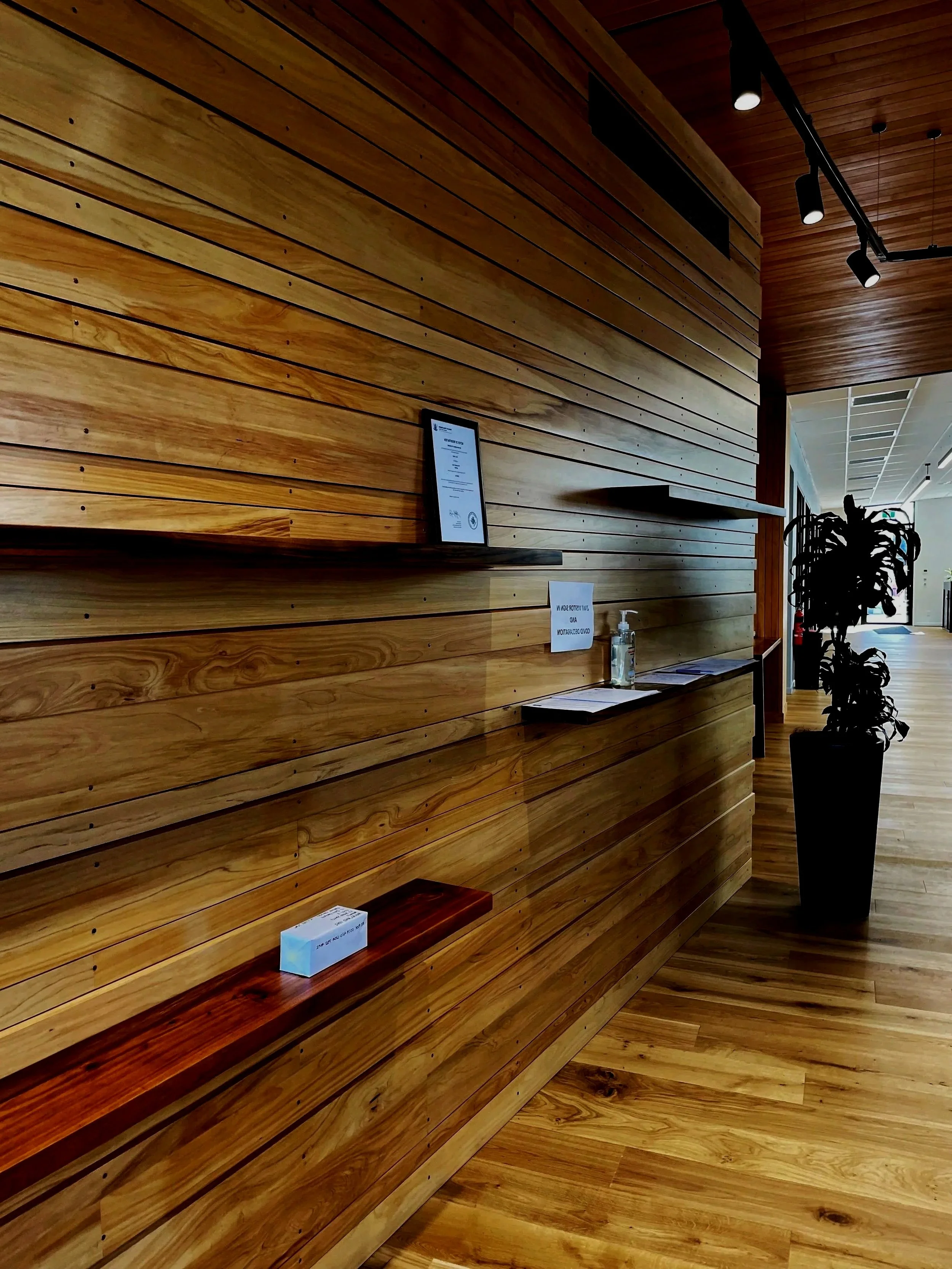 Wood-paneled wall with a small framed certificate, hand sanitizer, and small sign, next to a large potted plant in a hallway.