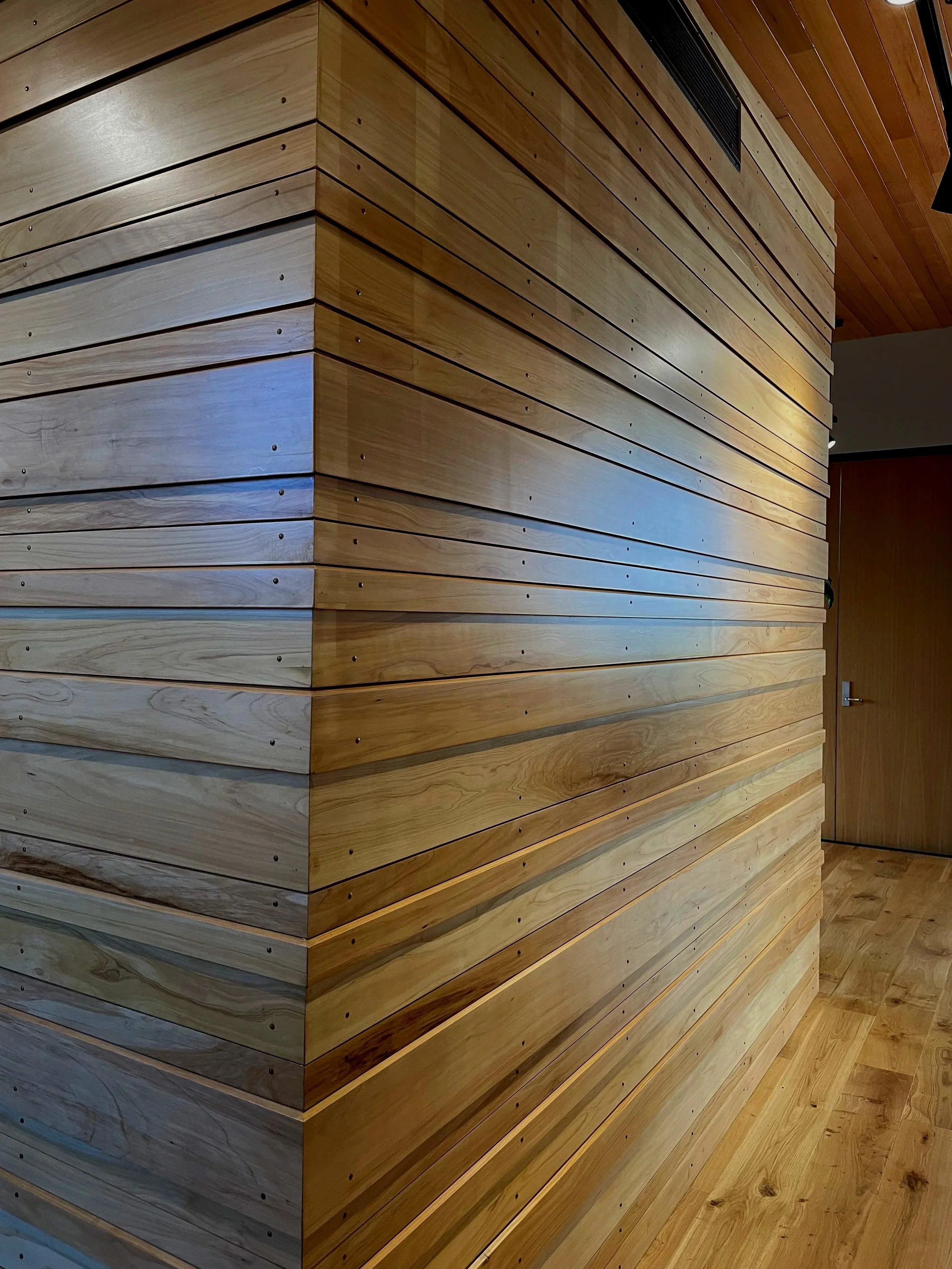 Corner of a room with hardwood paneling on the walls and ceiling, and a wooden floor, with a door visible on the right side.