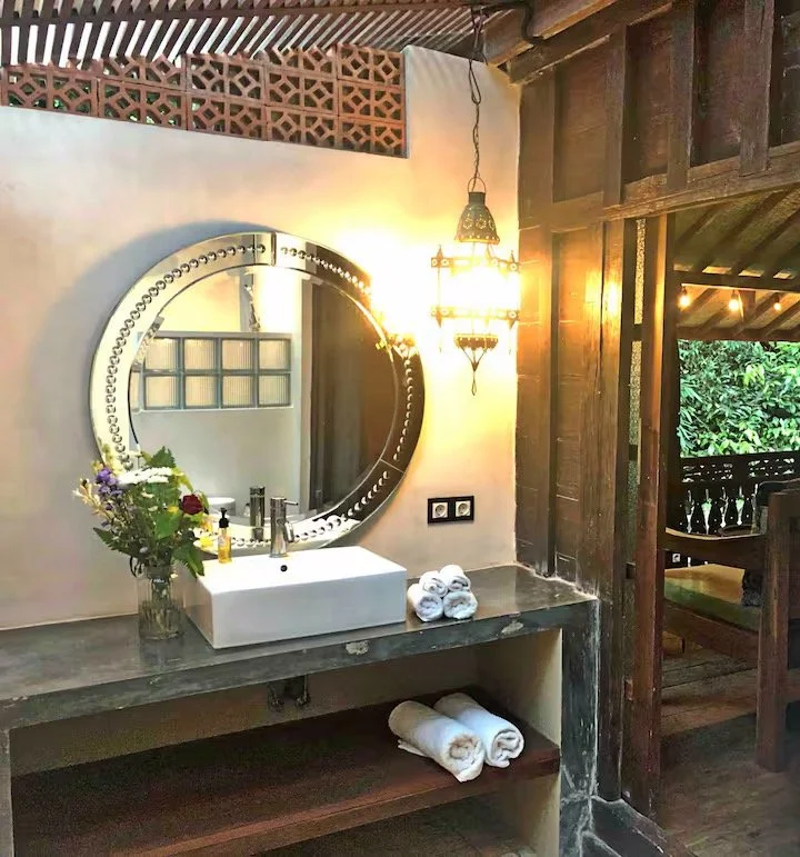 Bathroom sink area with a round mirror, a flower vase, rolled towels, and a Tiffany-style hanging lamp, with rustic wooden walls and outdoor view.