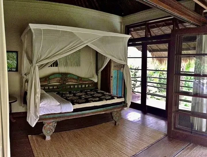 Room with a four-poster bed with cream curtains, wooden floor, and large glass doors leading outside to a lush green view.