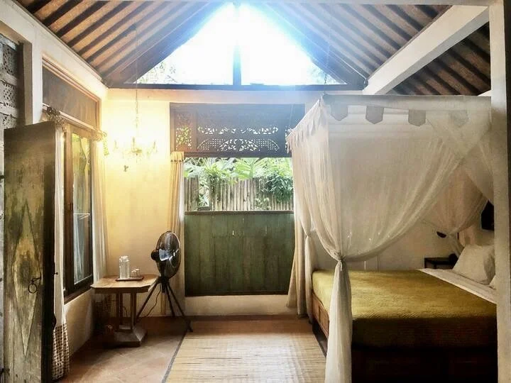 A cozy bedroom with a four-poster bed, mosquito net, and large window with bamboo fence view, natural sunlight, and wooden accents.