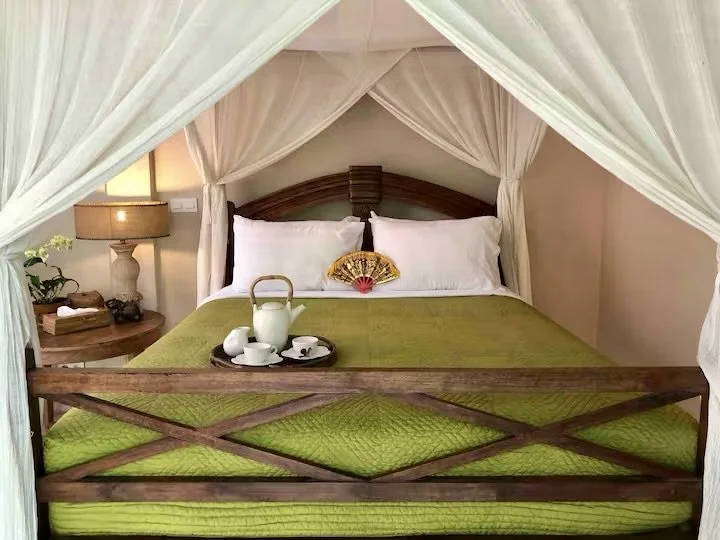 A cozy bedroom with a canopy bed, green bedding, and a tray holding a teapot and cups. Soft lighting from a bedside lamp enhances the warm atmosphere.