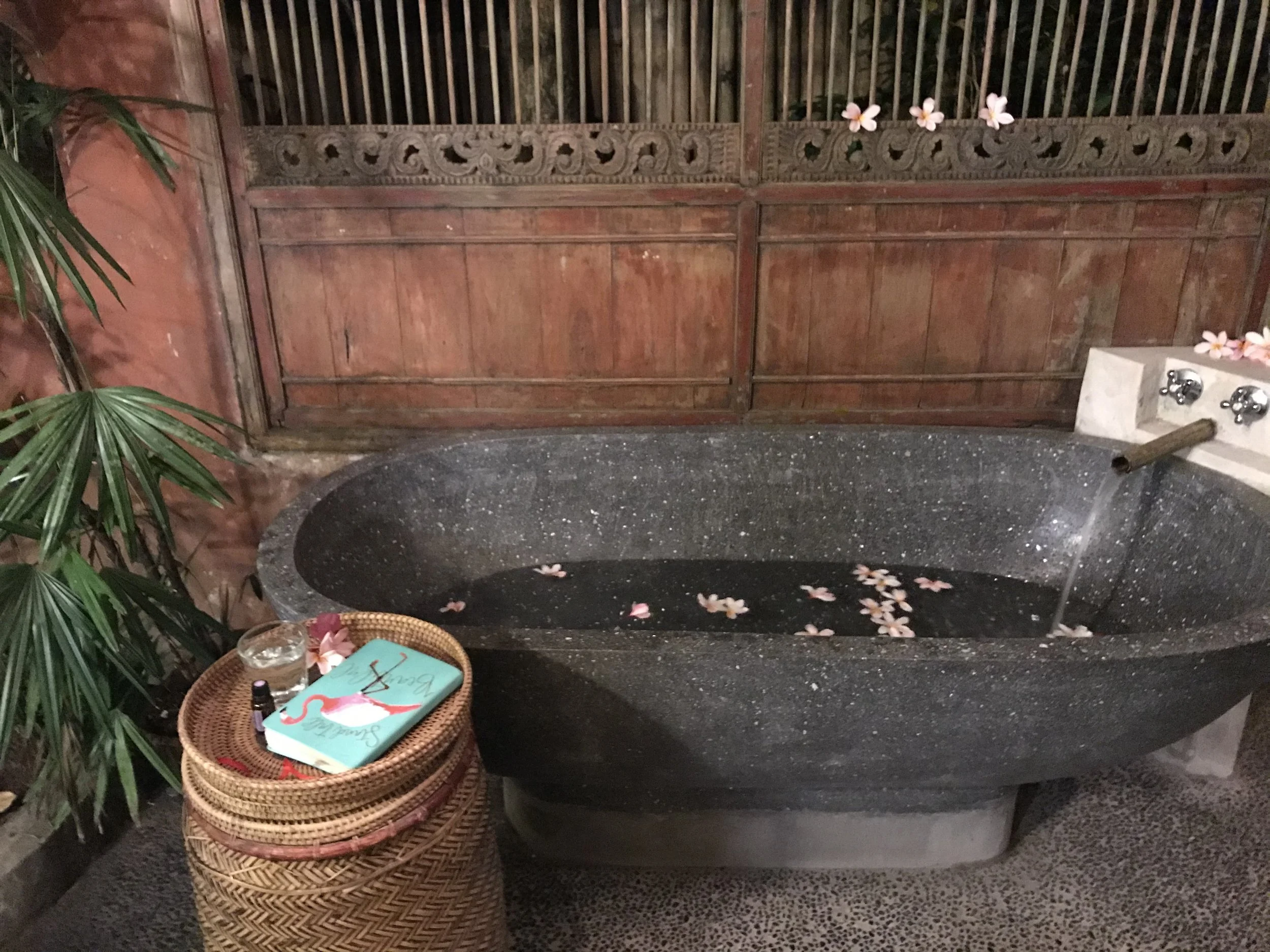 A black granite bathtub filled with water and floating pink flower petals, against a wooden Indonesian-style carved wall, with a wooden spout pouring water into the tub. To the left, a wicker basket holding a glass of water, a small bottle, and a tray with a teal-colored book or magazine and a pink flamingo illustration. There is also greenery on the left side.
