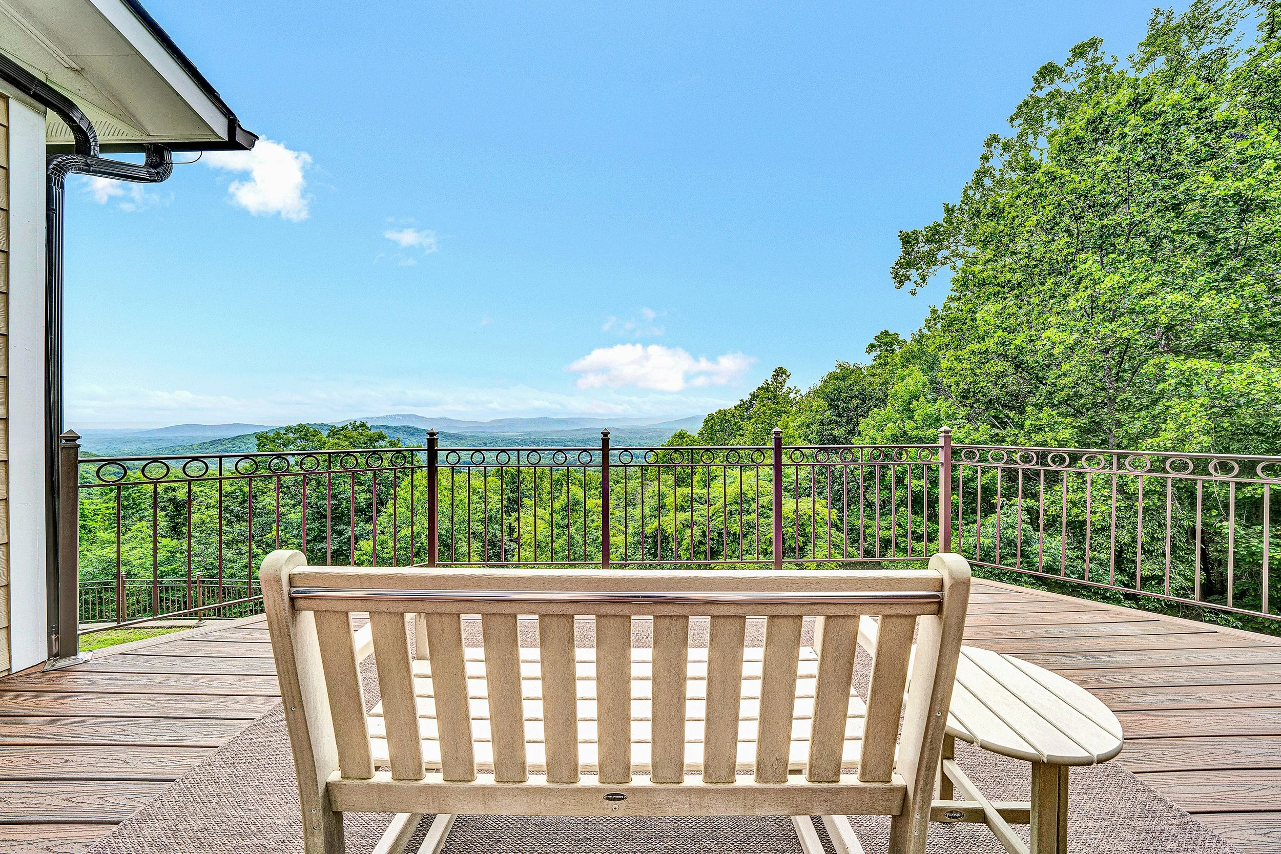 A view from a wooden deck with a bench, overlooking a lush green landscape with trees and distant mountains under a bright blue sky. Part of the house with beige siding and black gutters is visible on the left side.