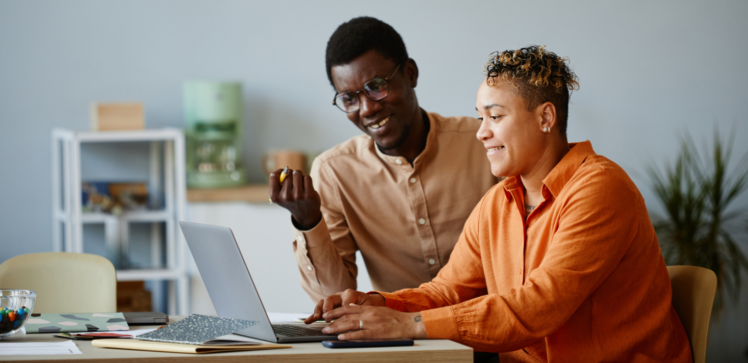 Two people working together at a desk with a laptop and papers in a bright room, laughing and engaging.