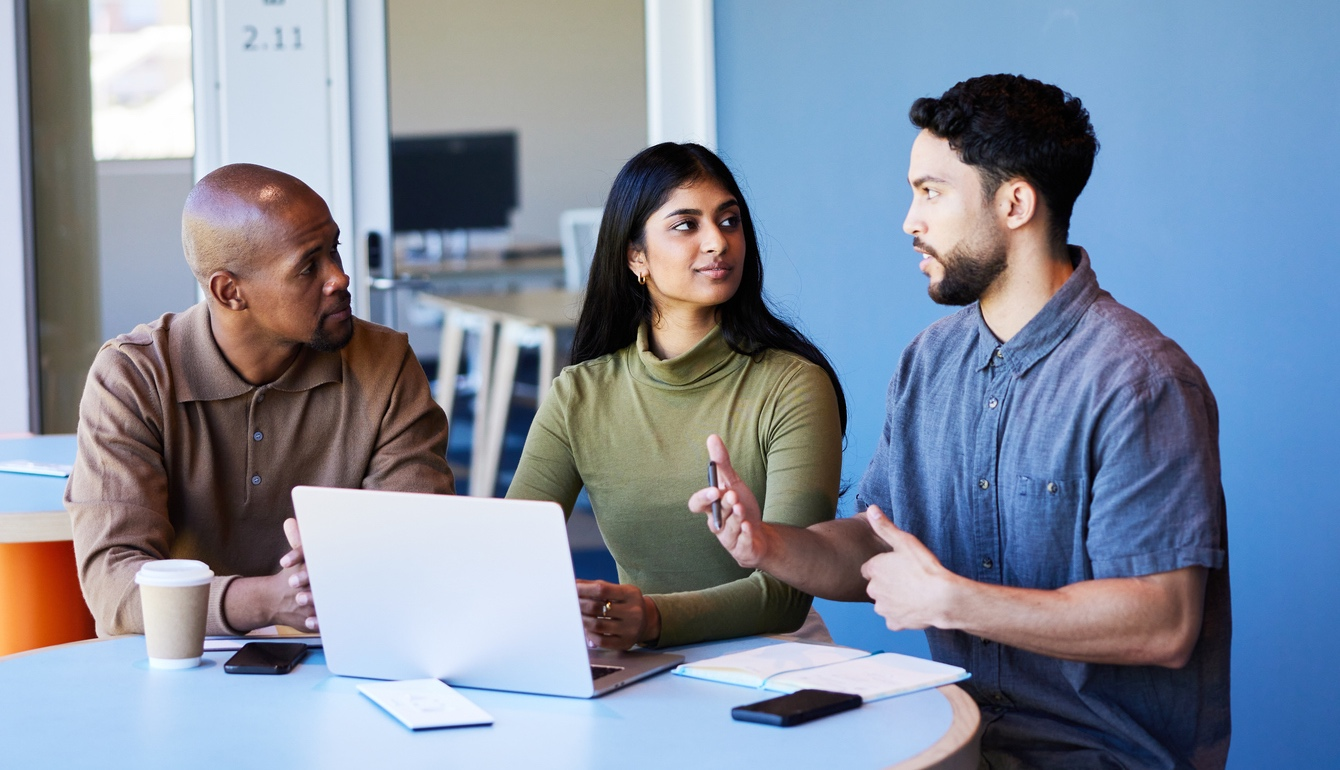 Three people having a discussion at a table in an office. The man on the right is speaking, the woman in the middle is listening, and the man on the left is looking at the speaker. There is a laptop, a smartphone, a coffee cup, and notebooks on the table.