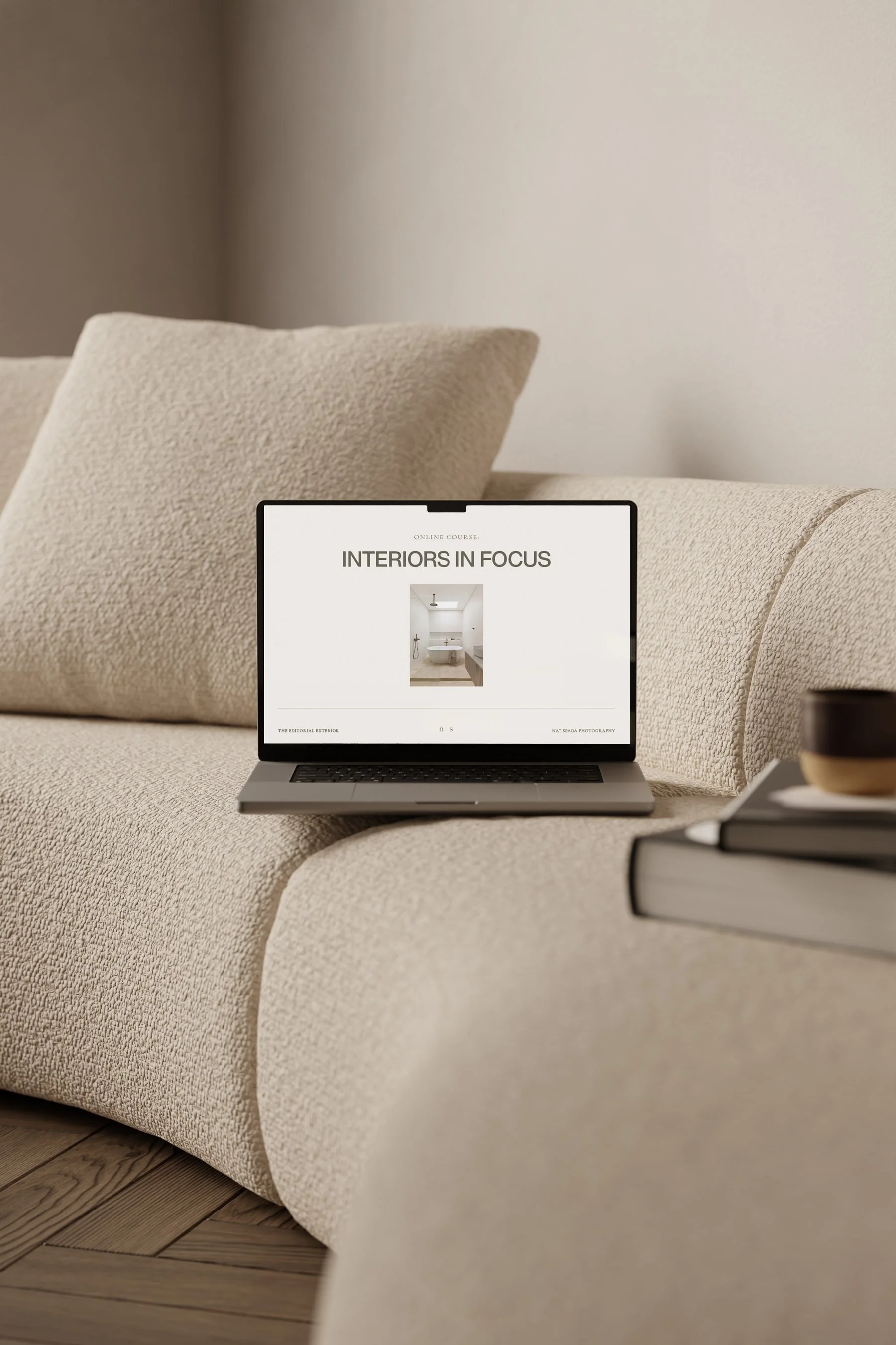 Laptop on a beige textured couch displaying an online course titled 'Interiors in Focus' with a bathroom interior image, next to a stack of books and a black bowl on the right.