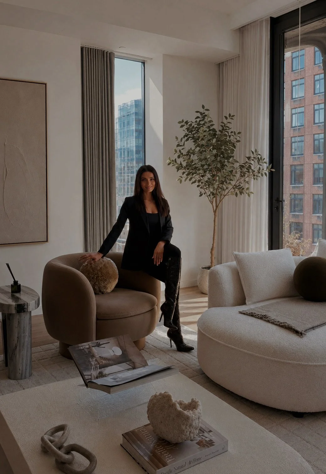 A woman in black clothes standing in a modern living room with large windows, a potted plant, beige and white furniture, and decorative objects.