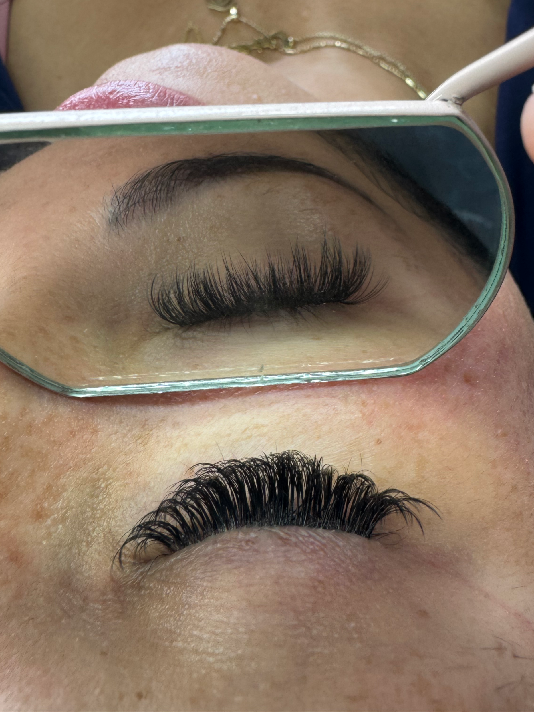 Close-up of a person's eye with eyelash extensions being applied, seen through a mirror reflecting the eye and eyelash technician's hand