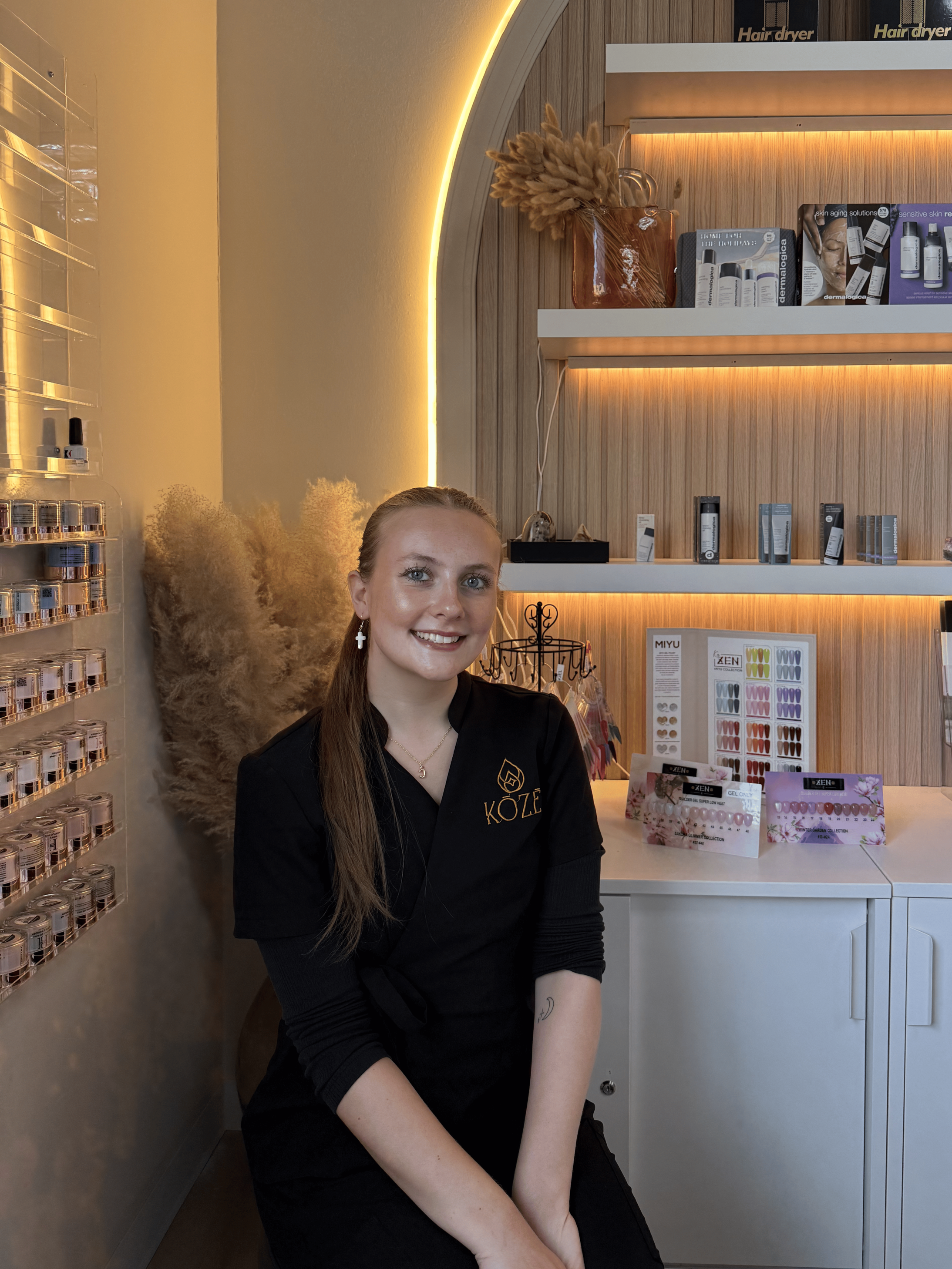 A woman sitting in a spa or salon, wearing a black uniform with a name badge. Behind her is a wall with shelves containing skincare products and a black hair dryer box. To her left is a tall vase with pampas grass illuminated by decorative lighting.