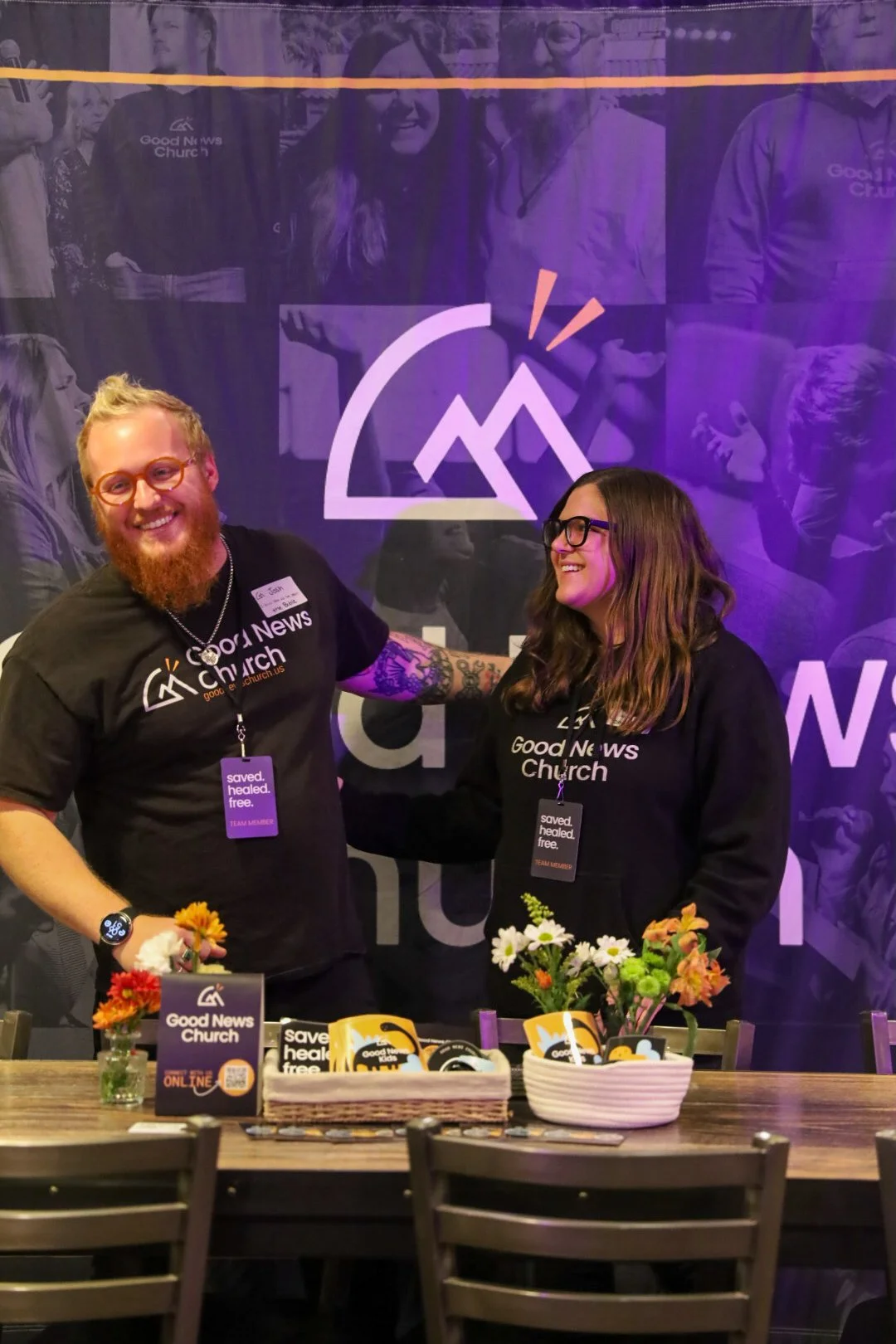 Two smiling people, a man with a beard and glasses, and a woman with glasses, standing behind a table with flowers and church merchandise, in front of a purple and black backdrop with the Good News Church logo.