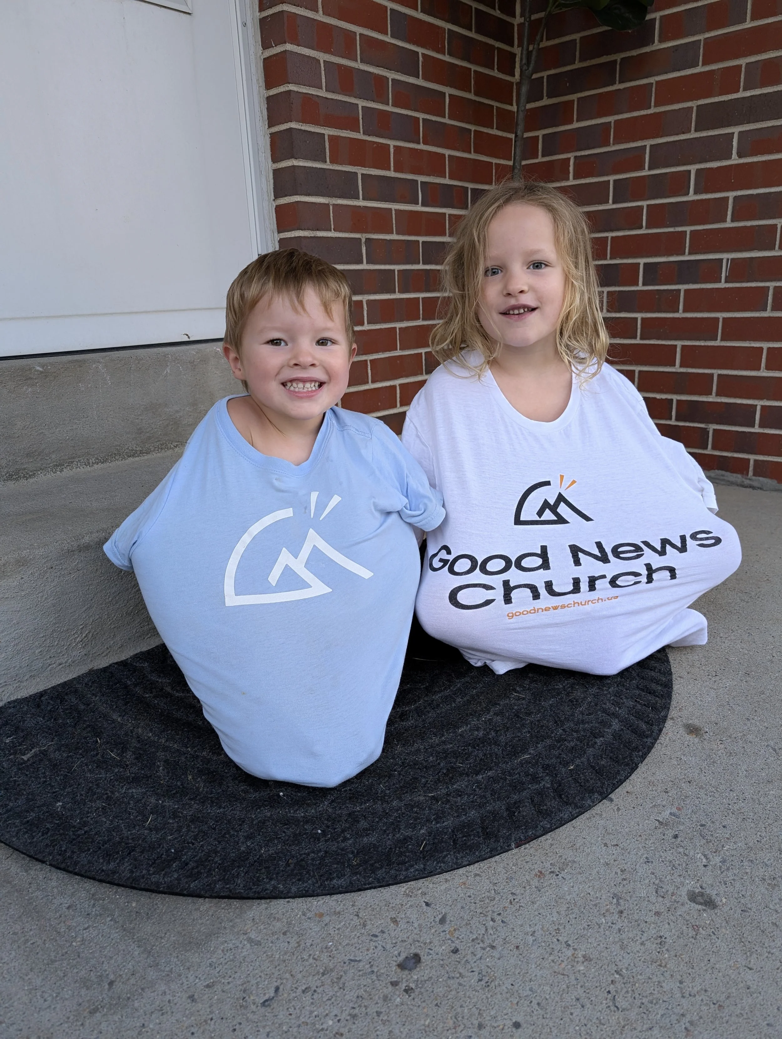 Two children sitting on a mat outside a brick building, wearing t-shirts with a church logo and text reading "Good News Church"