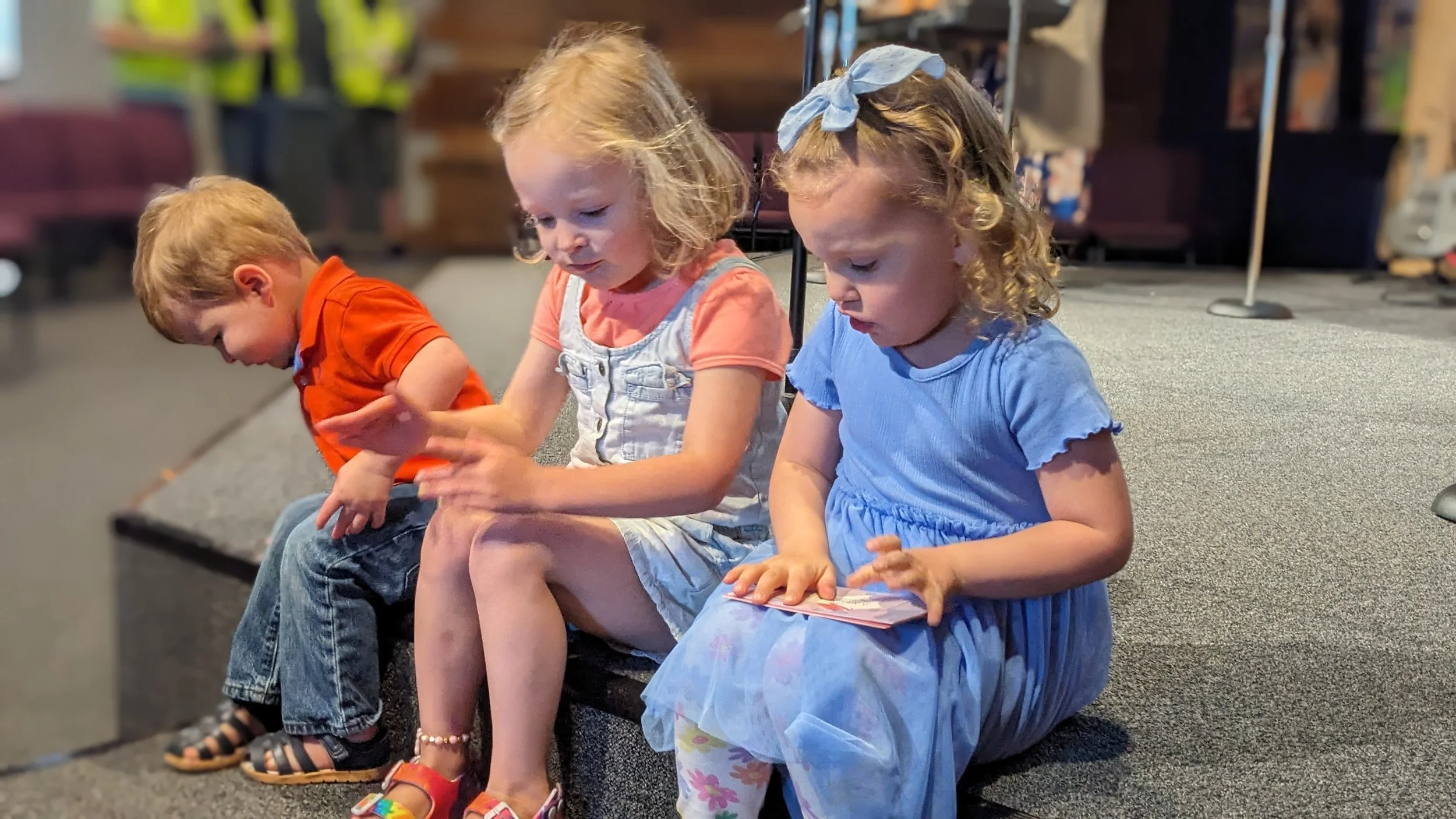 Three young children sitting on a stage or platform, looking down at objects in their hands, with a blurry background of a room or auditorium.