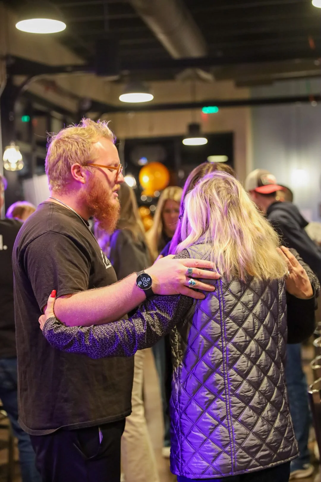 People in a dimly lit social setting, engaging in supportive or comforting gestures.