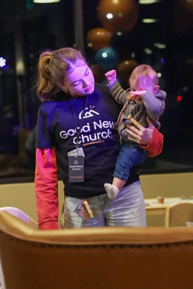 A young woman wearing a 'Good News Church' t-shirt holds a crying toddler in her arms, with balloons in the background at night.