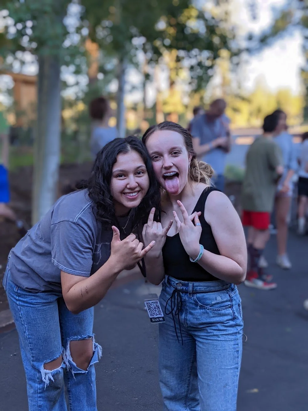 Two young women smiling and making hand signs at an outdoor gathering, with other people in the background. The woman on the right is sticking out her tongue.