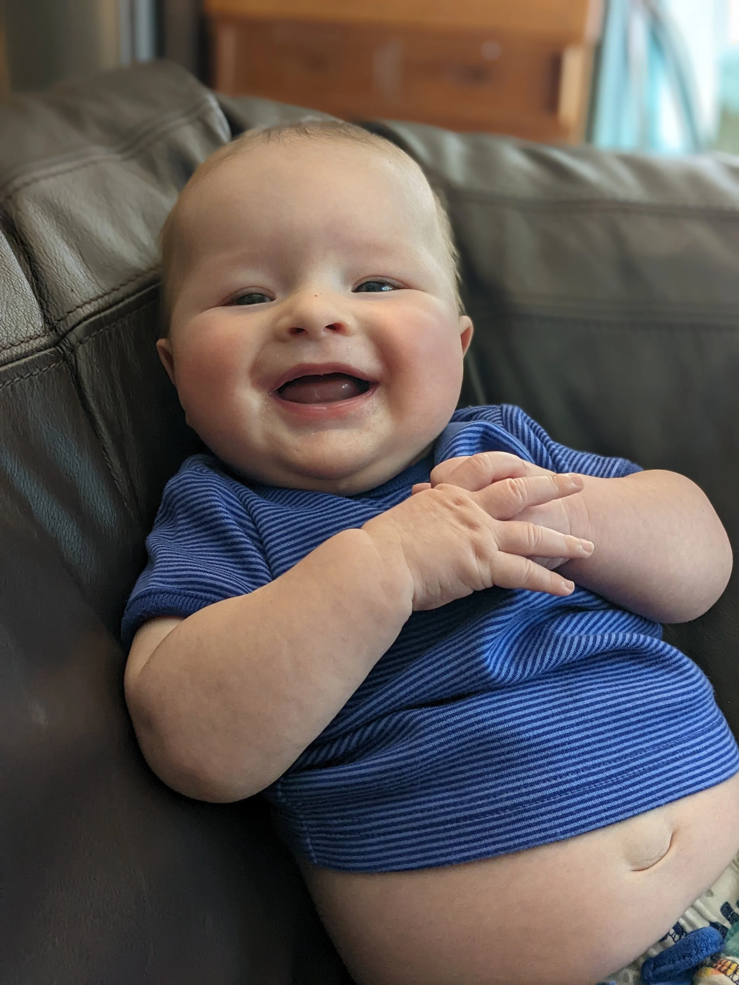 Smiling baby boy with blue eyes and wearing a blue striped shirt sitting on a leather couch.