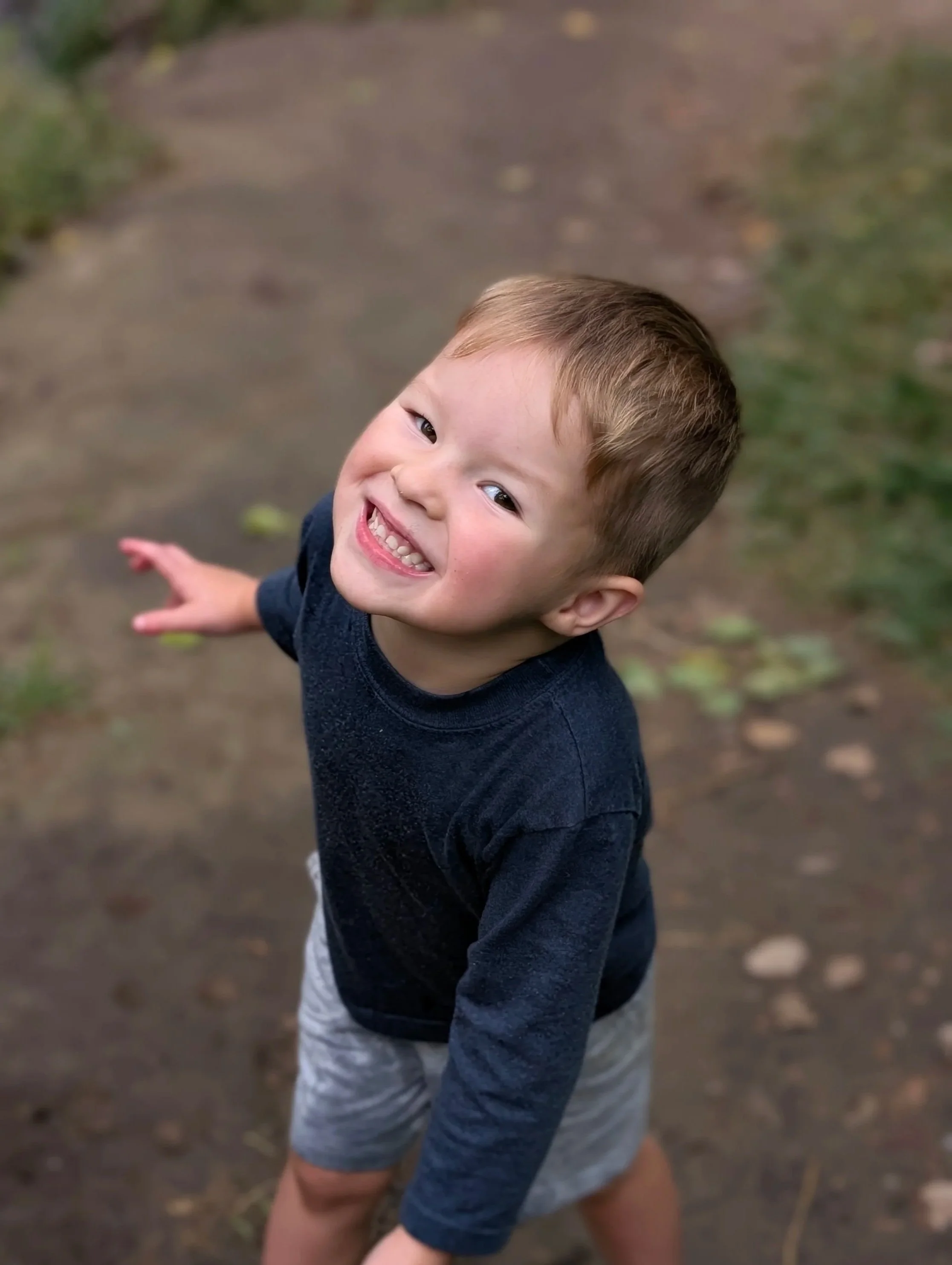Happy young boy smiling and looking up while outside on a dirt path surrounded by greenery.