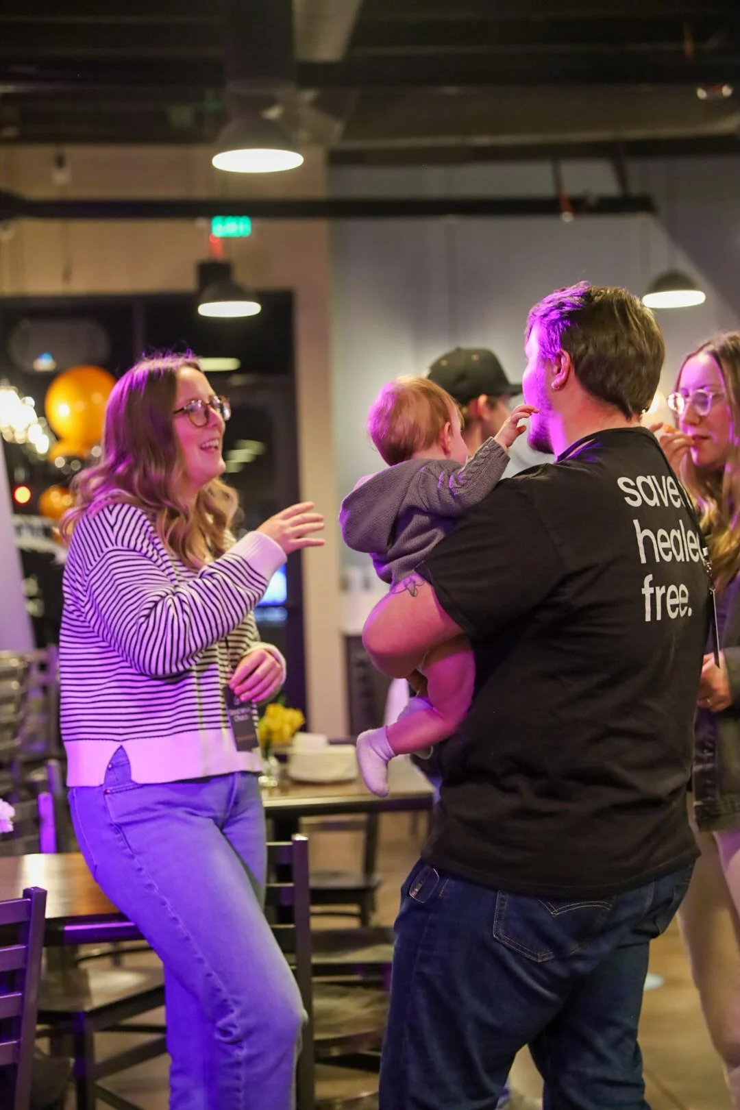 A group of four people, including a man holding a child, are interacting and smiling inside a restaurant or cafe. The man is wearing a black T-shirt with white text, and the child is reaching towards his face. The woman on the left, with glasses and wavy hair, is wearing a striped sweater and jeans, and is engaging with the group. The setting features dim lighting and a casual atmosphere.