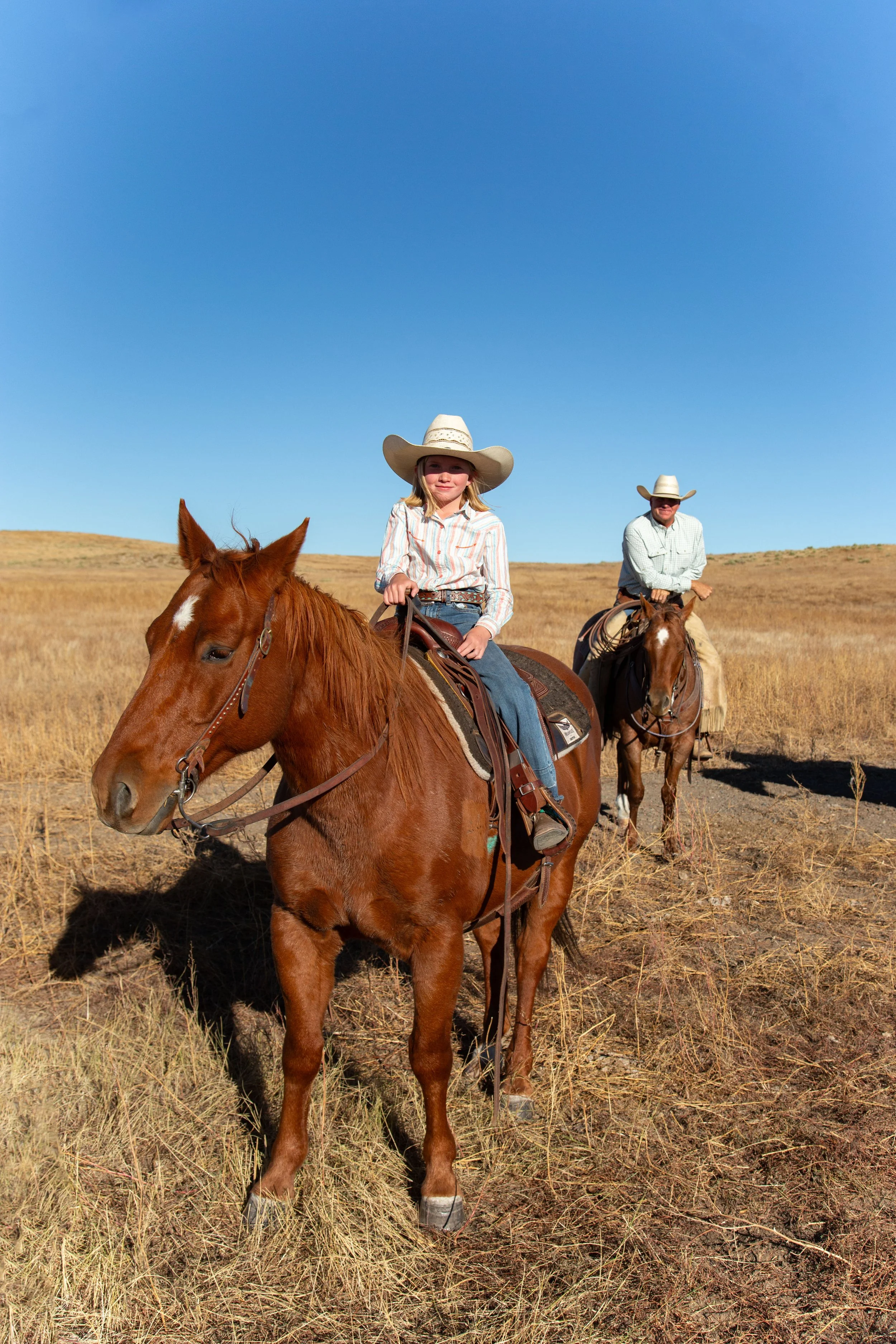 A young girl and an adult man riding horses in a dry, open field with a clear blue sky.