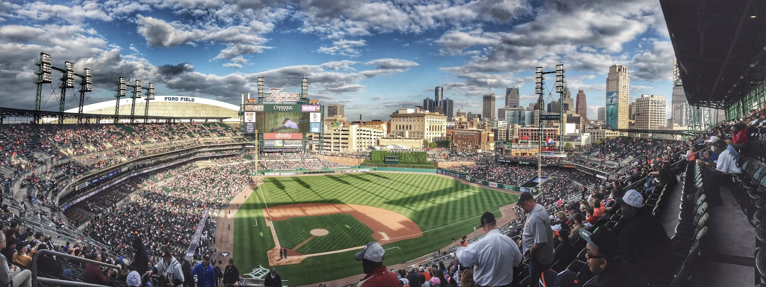 Wide-angle view of a baseball stadium with a partially filled seated crowd, green field, and skyline of tall buildings in the background on a partly cloudy day.