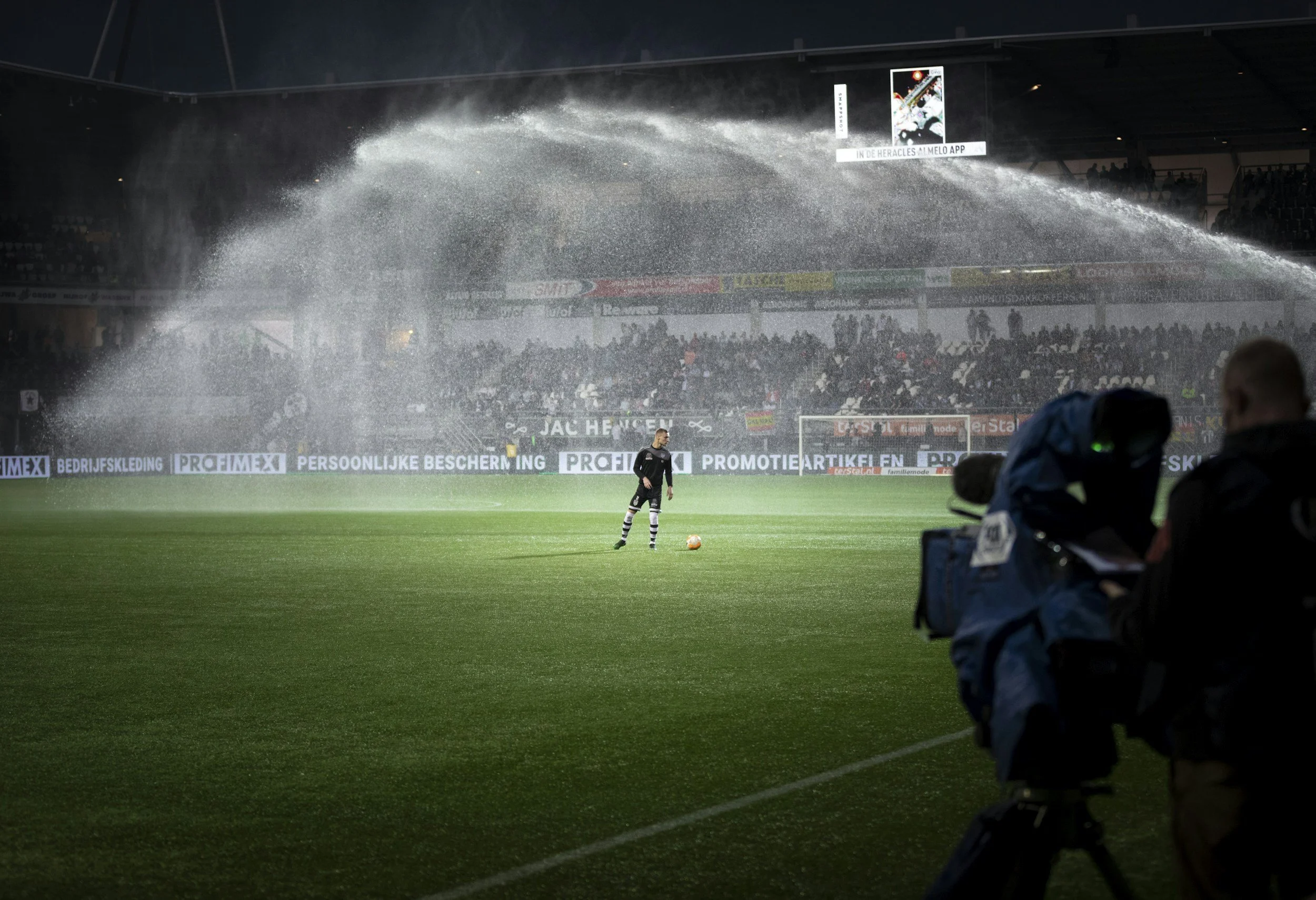 A soccer player standing on a rain-soaked field with a ball, with water being sprayed overhead by sprinklers in a stadium filled with spectators and a cameraman in the foreground.