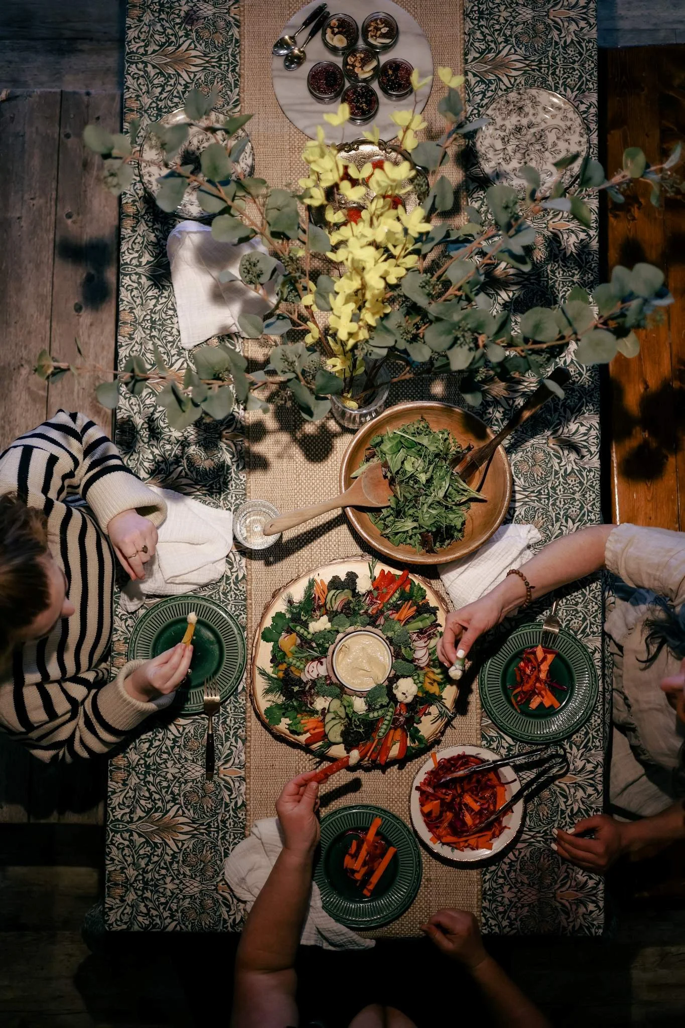 Top-down view of a dinner table with various dishes, salads, small bowls of sauce, and a large vase with yellow and green foliage, set on a patterned tablecloth with some wooden floors visible.