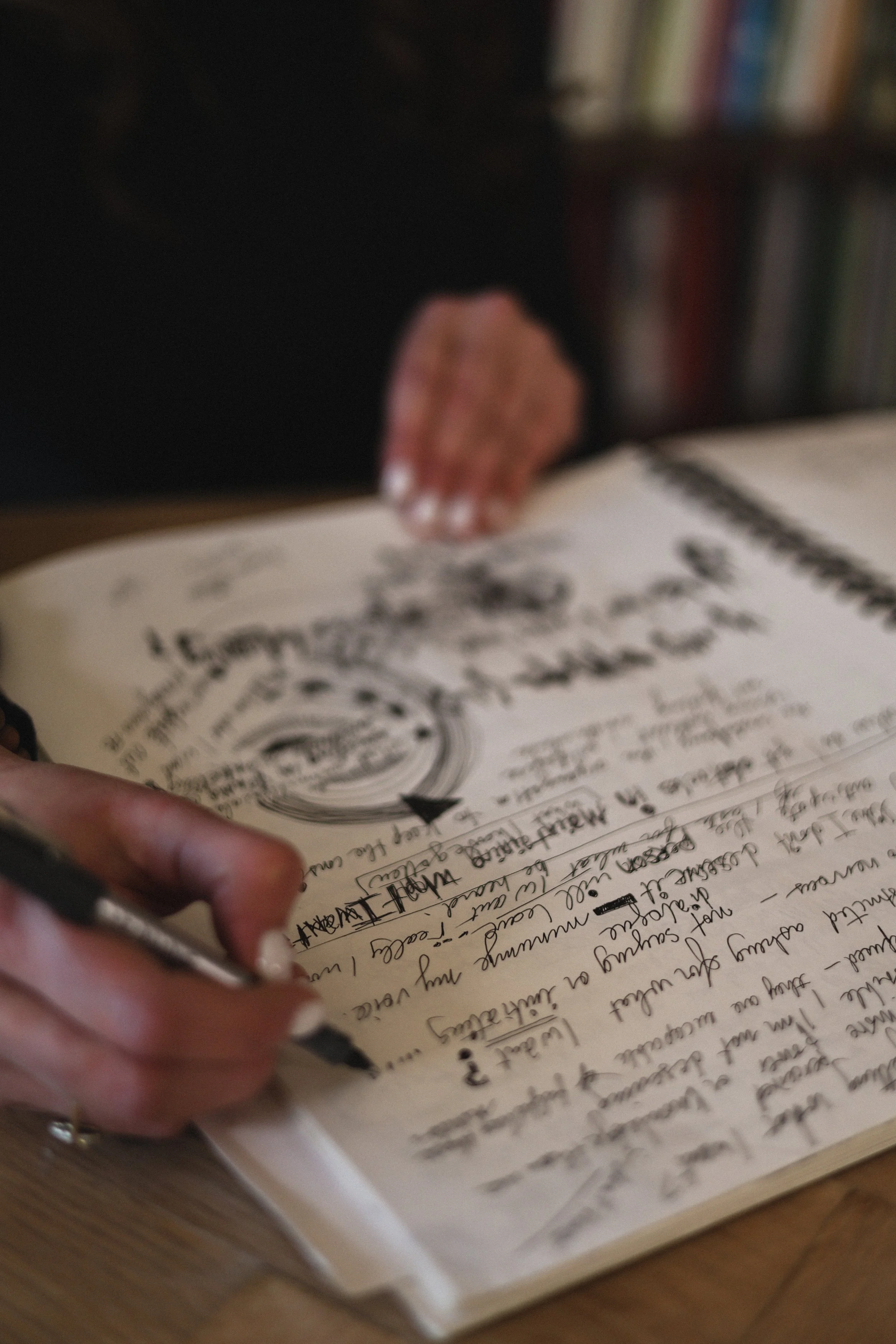 Close-up of a person's hand writing notes in a notebook with cursive handwriting and a pen, resting on a wooden surface.