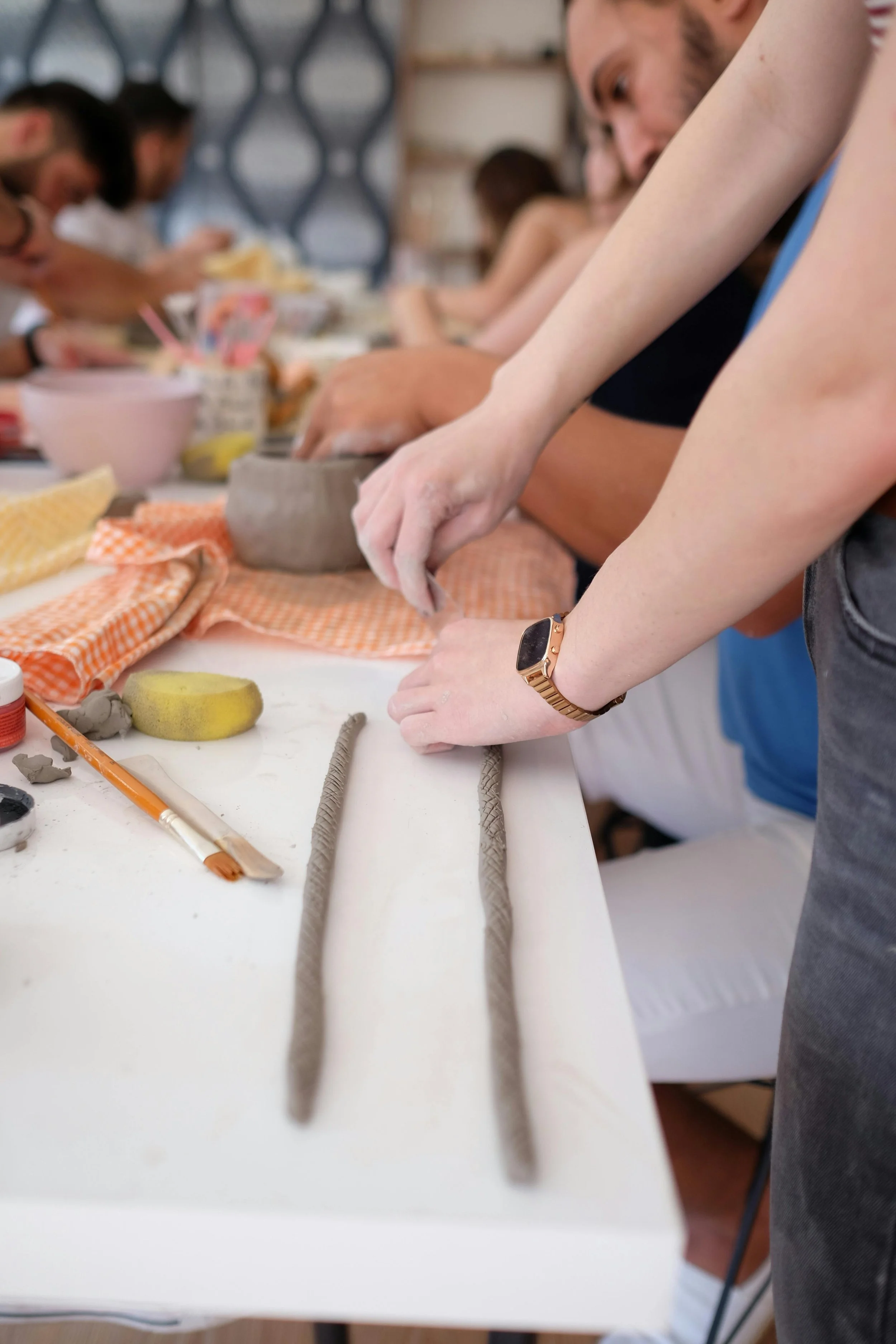 People participating in a pottery or clay-making workshop, working with clay and tools on a long white table.