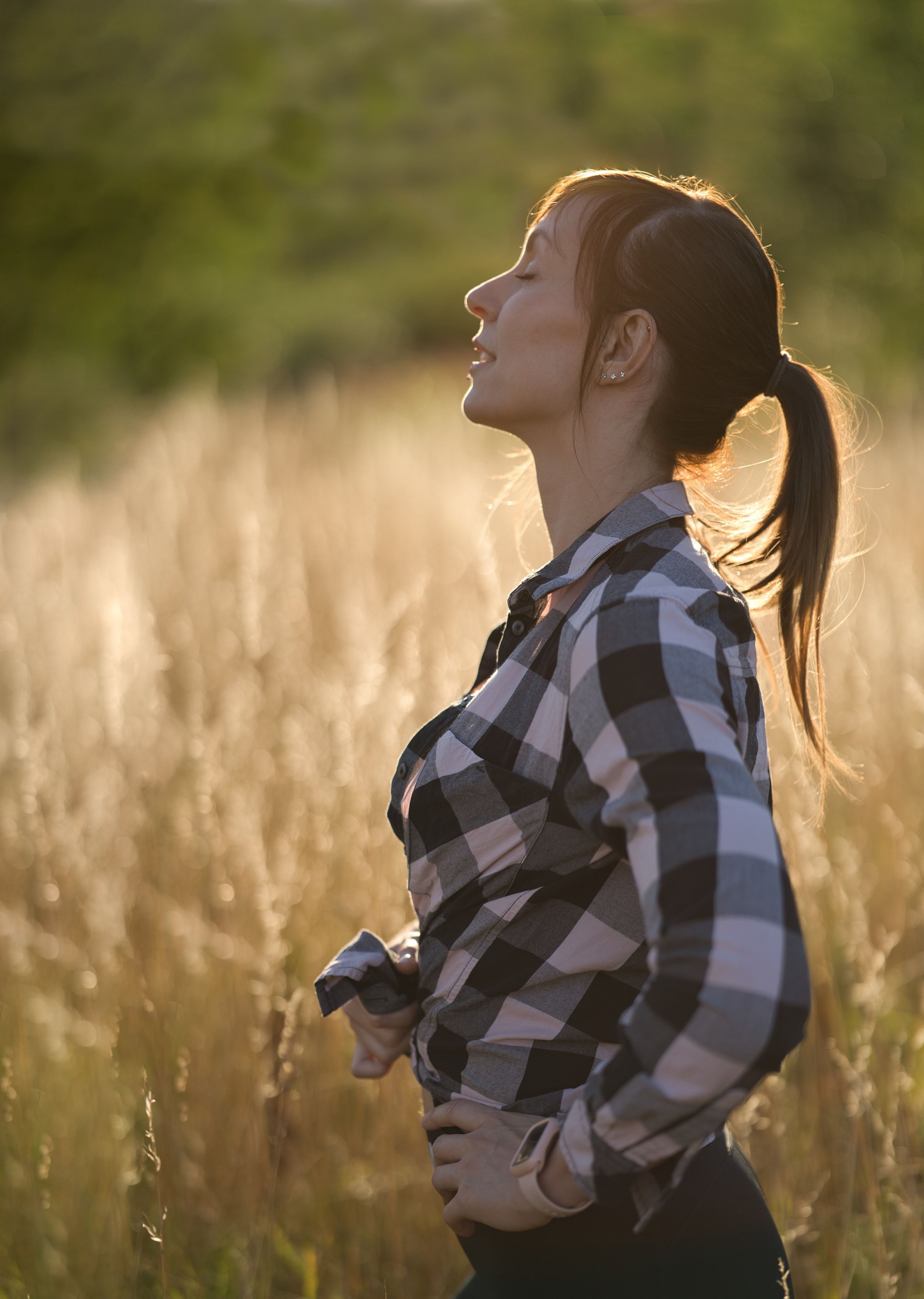 A woman with brown hair tied in a ponytail, wearing earrings and a black and white checkered shirt, standing in a field of tall, golden grass during sunset, with her eyes closed and a peaceful expression.