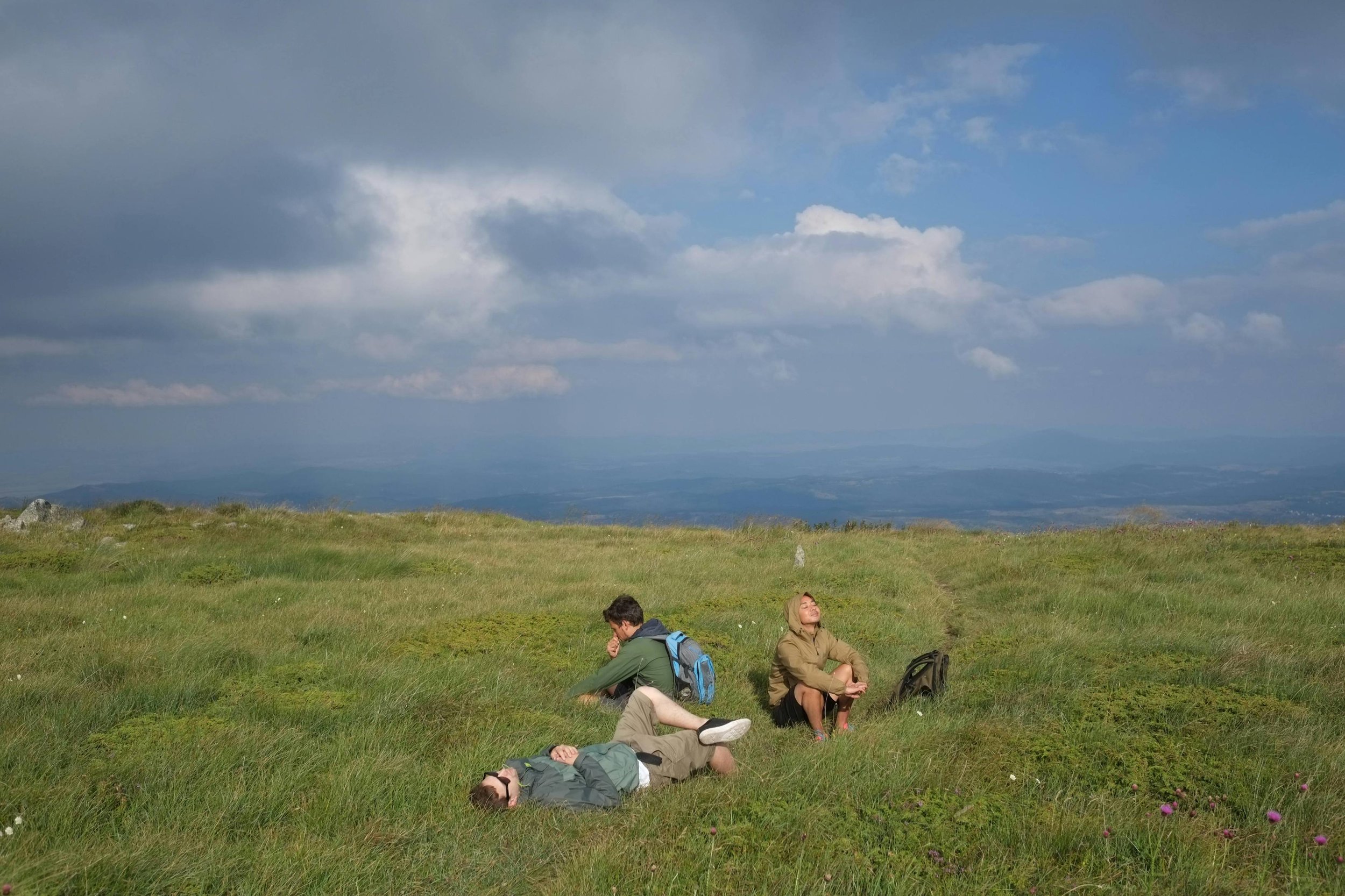 A group of hikers resting on a grassy hilltop with backpacks, some lying down and one sitting with eyes closed, under a partly cloudy sky with distant mountains in the background.