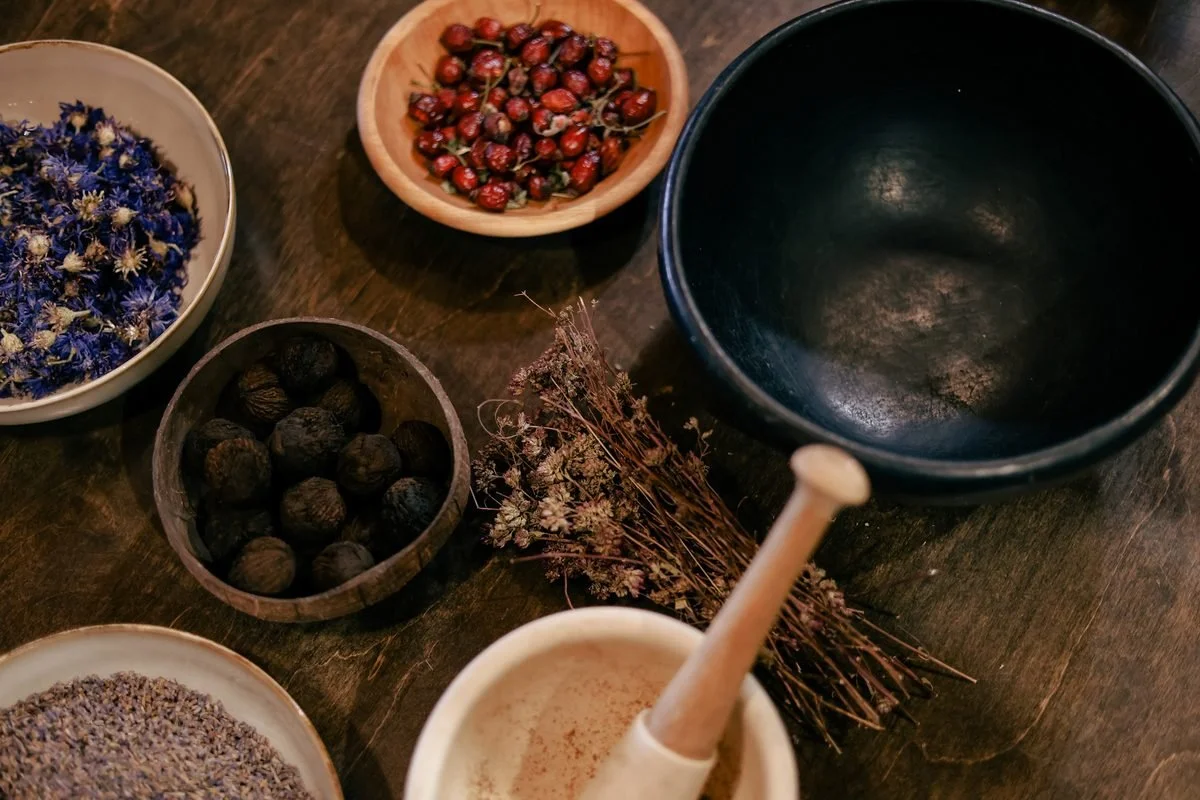 Dried herbs, rosehips, lavender, and blue flowers in bowls next to a mortar and pestle on a wooden table