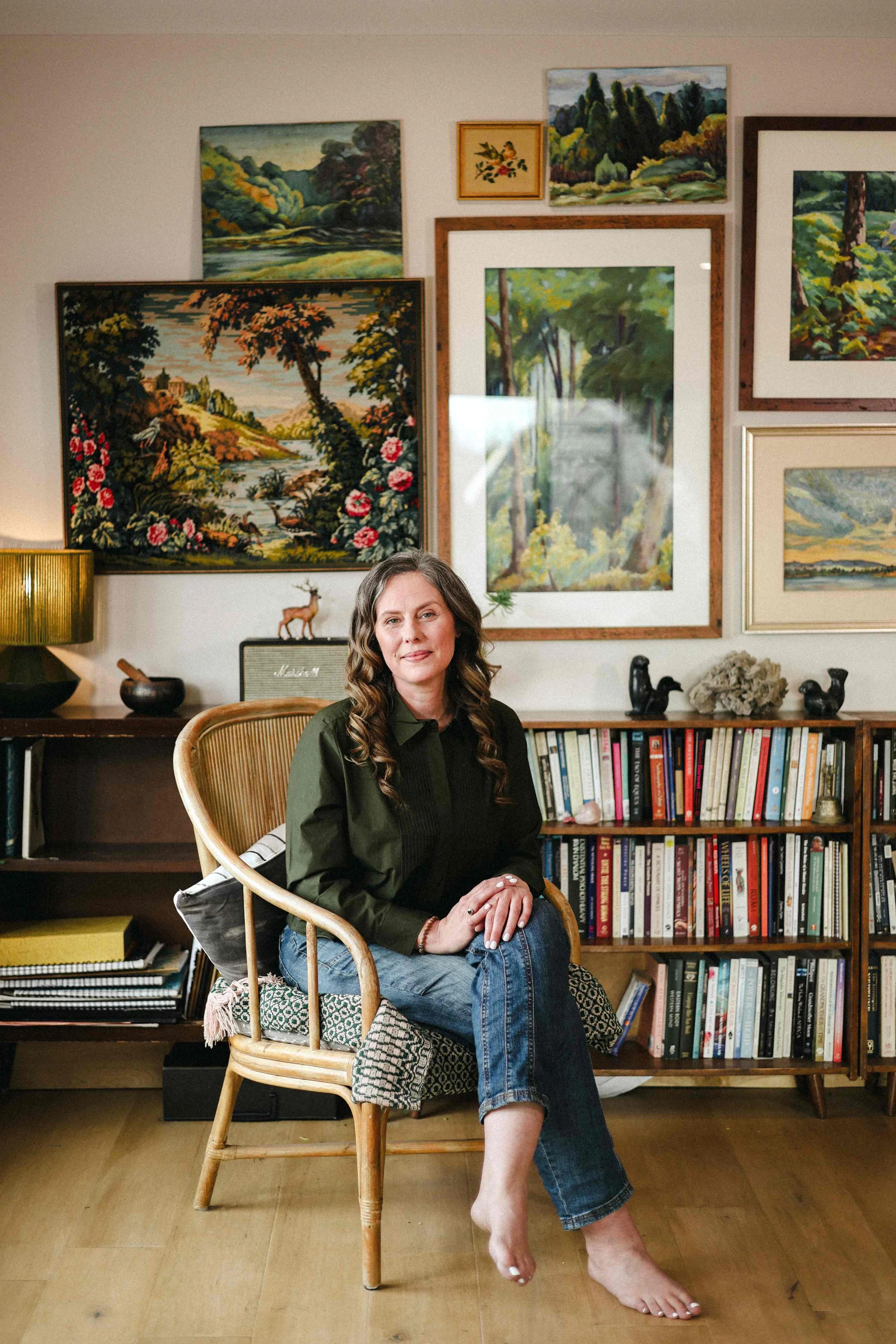 A woman with long brown hair and a green blouse sitting on a wooden chair with a patterned cushion in a room decorated with multiple landscape paintings and a bookshelf filled with books.