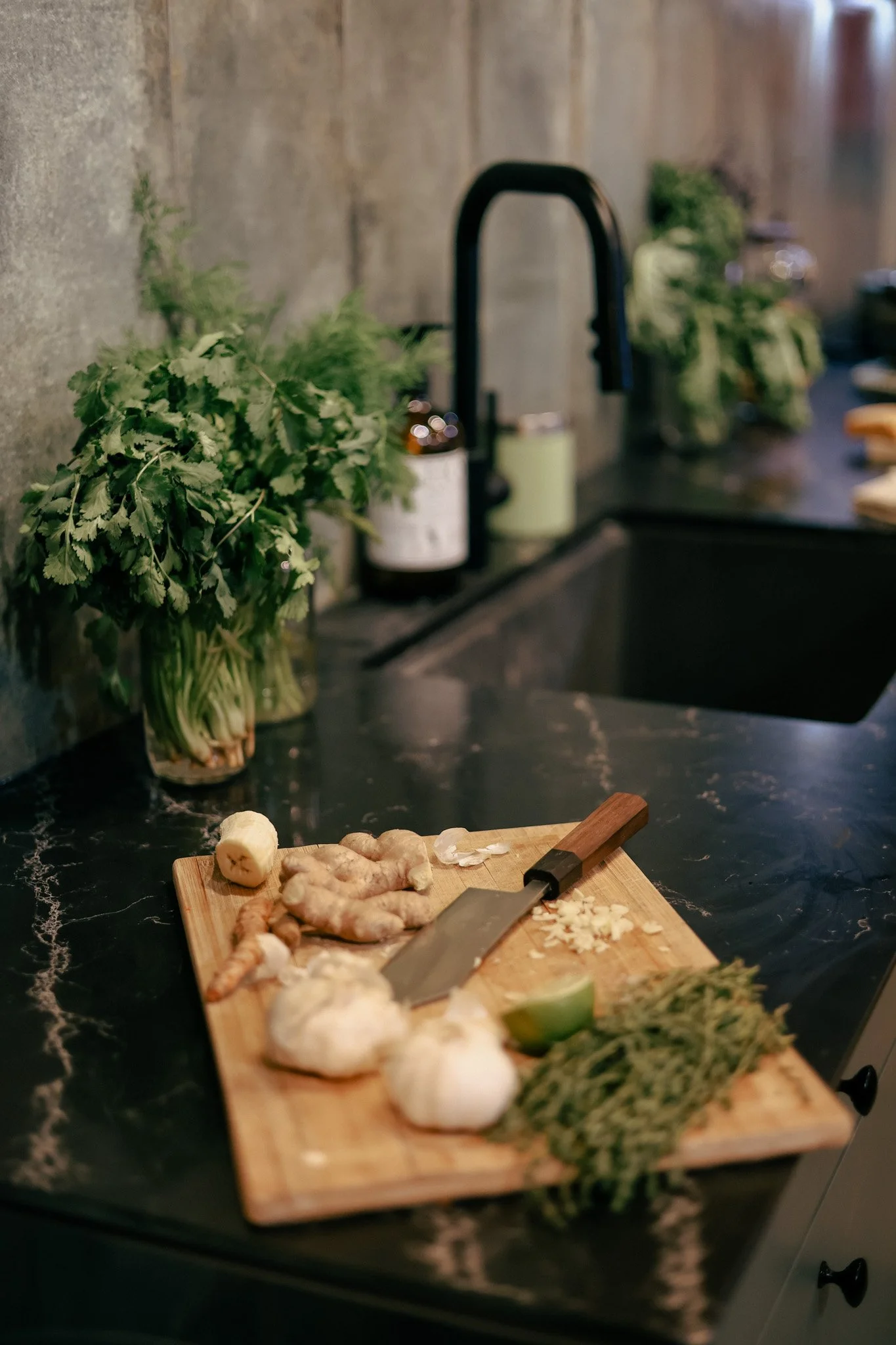 A wooden cutting board on a black marble countertop with chopped ginger, lime, and herbs, and a knife. In the background, there are fresh herbs, a sink with a black faucet, and blurred kitchen items.