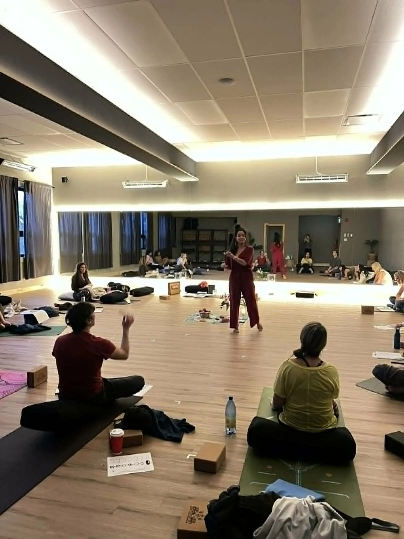 A group of people participating in a yoga or meditation class in a spacious, well-lit room with wood flooring and large windows with curtains. They are sitting on mats or cushions with some personal belongings and water bottles nearby.