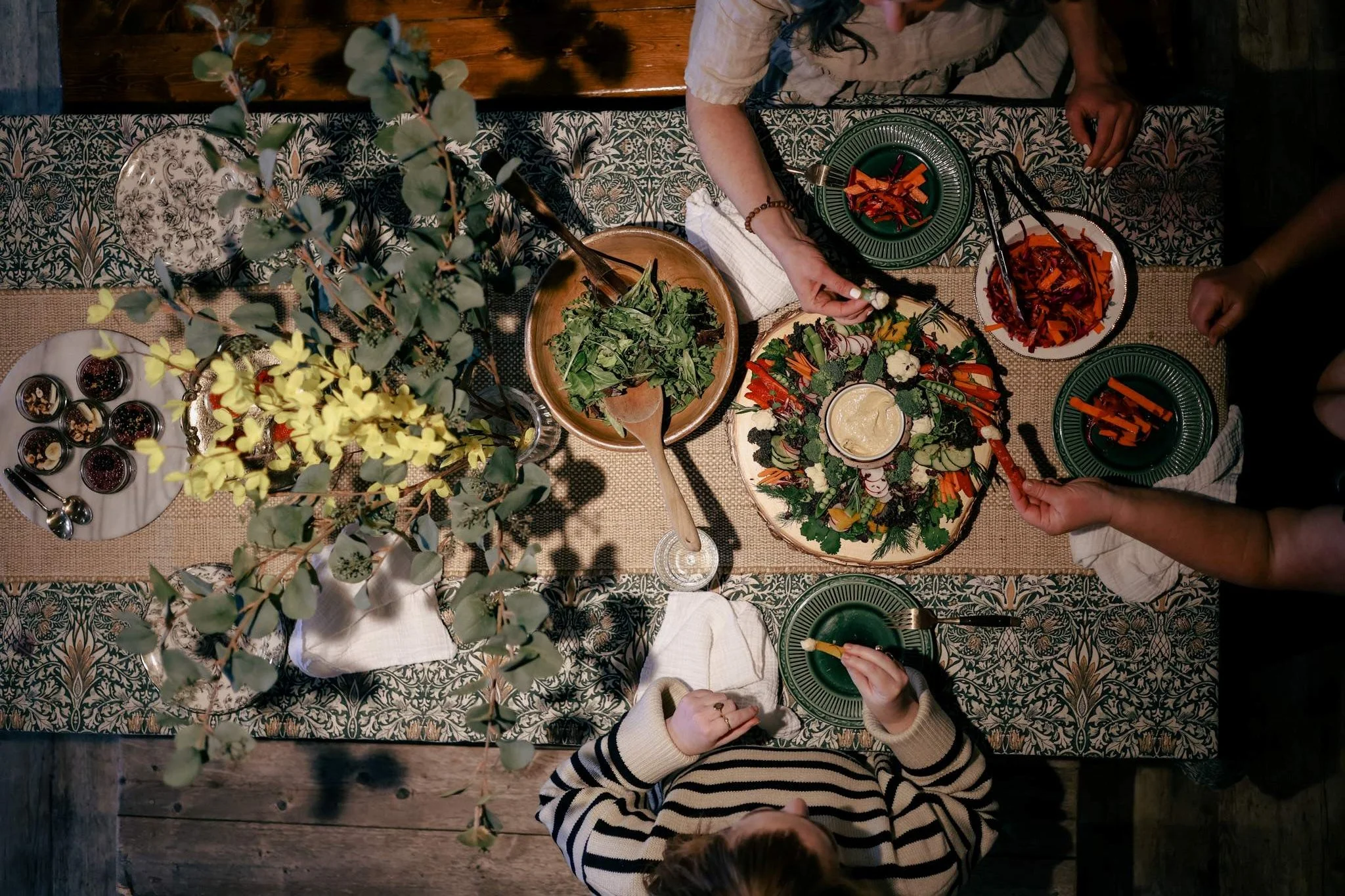 People sitting around a dining table topped with leafy greens, shredded vegetables, and a bowl of dip, sharing a meal.