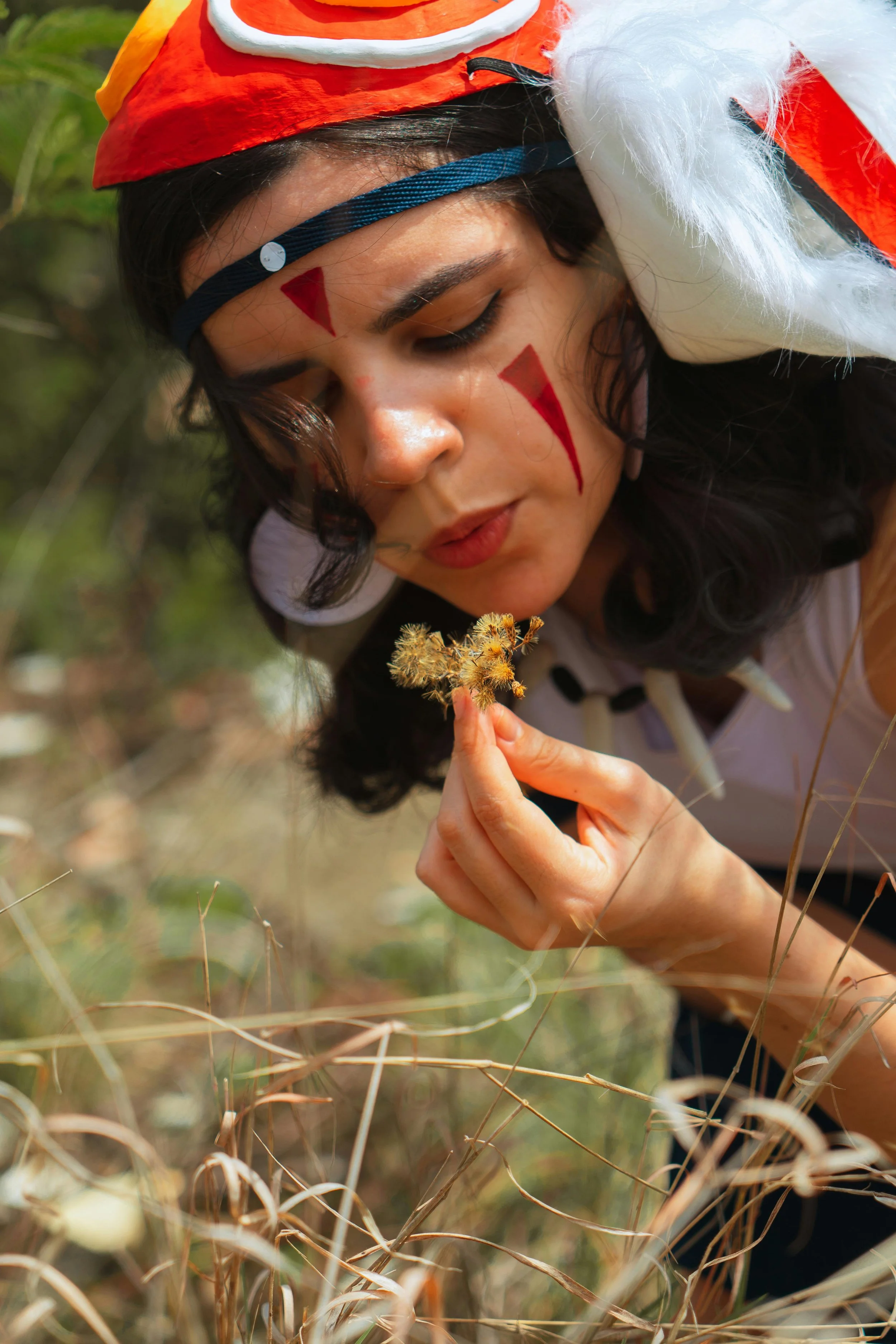 A person dressed in a costume with red and white face paint and a dragon hat, holding a small dried plant in their hand outdoors among dry grass.