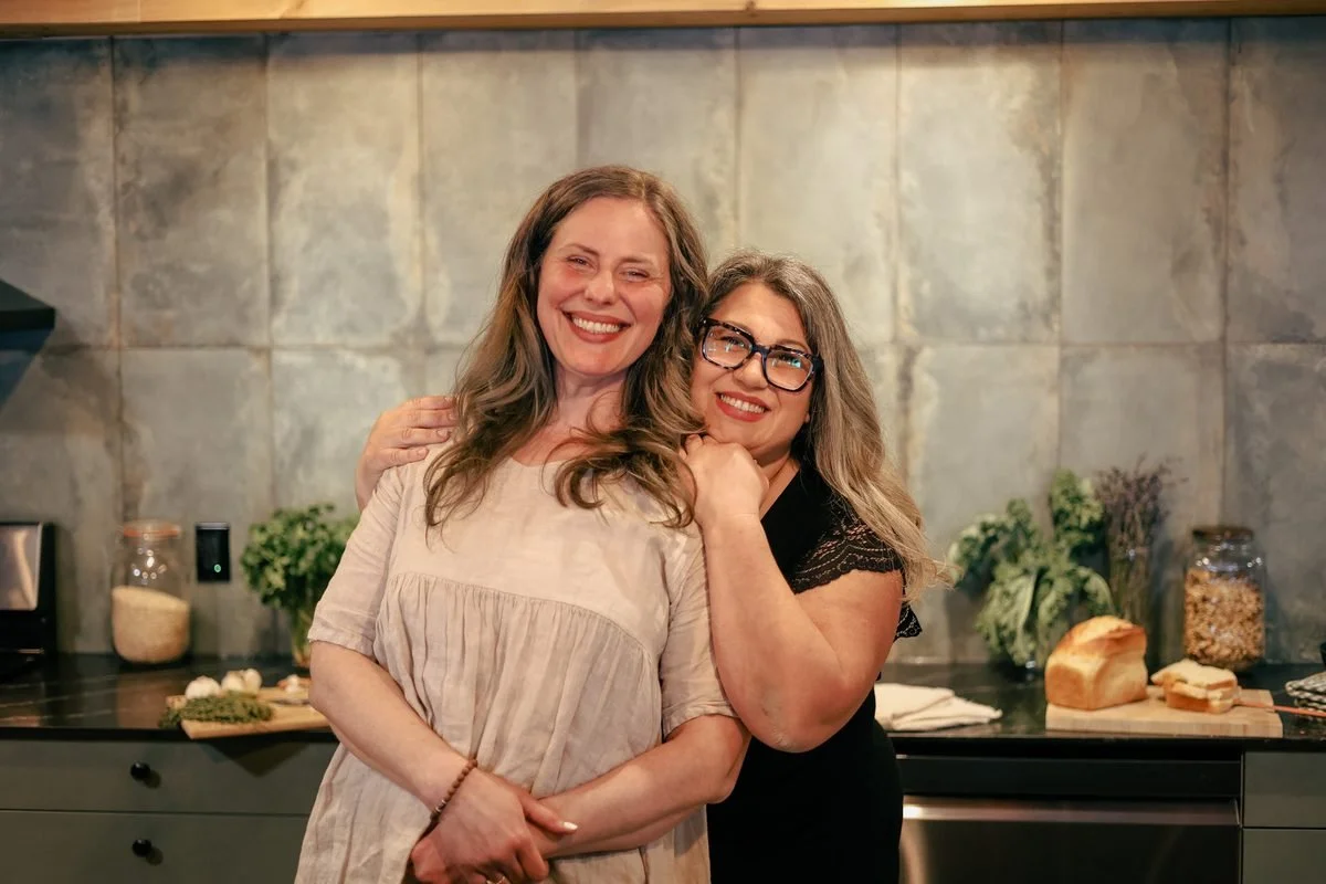 Two female chefs and hosts smiling together in a rustic kitchen next to freshly baked bread and herbs
