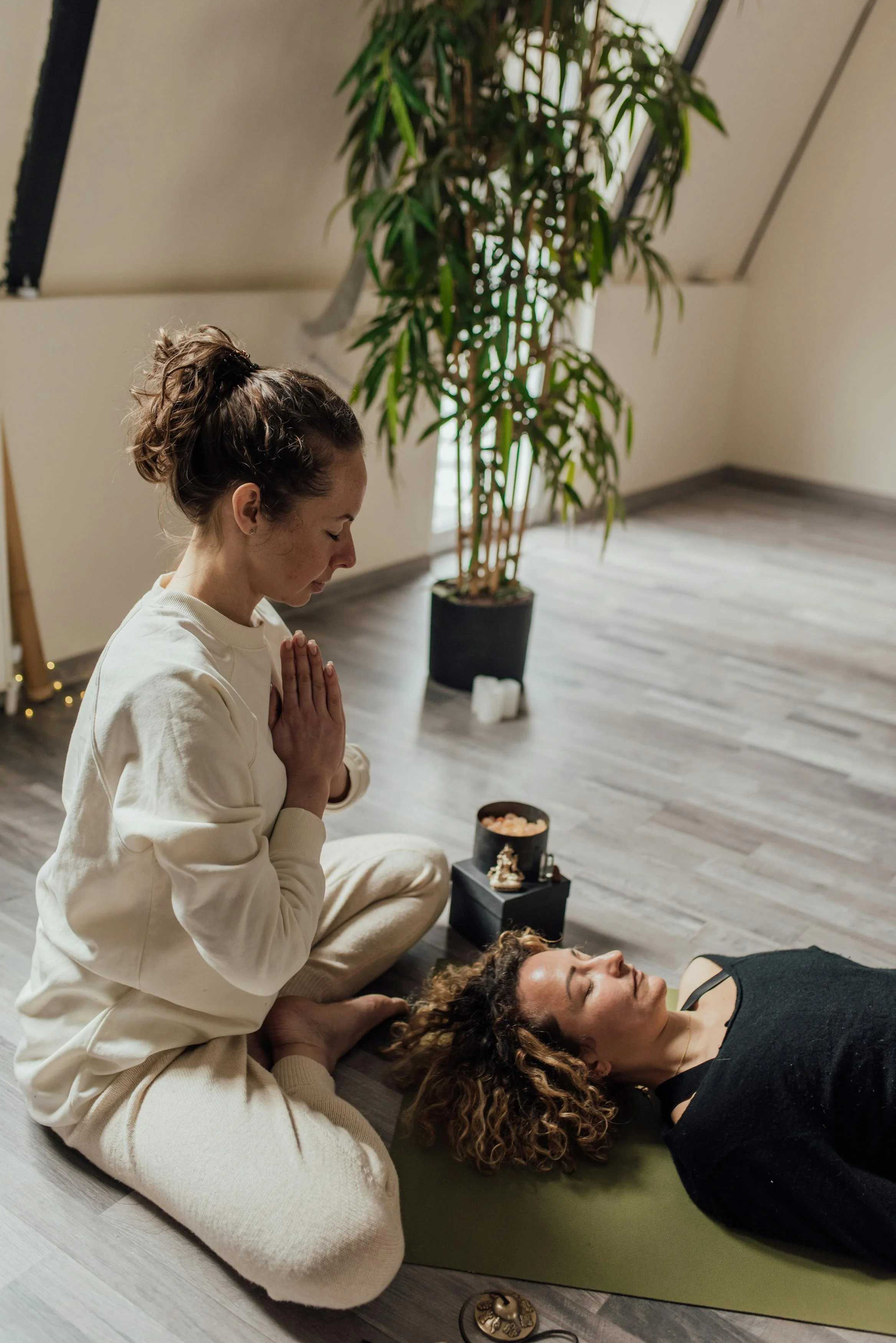 A woman is practicing yoga or meditation, sitting cross-legged with her hands in a prayer position, eyes closed, in a room with a large potted plant and candles. Another woman is lying down on a yoga mat, relaxed, with a therapist or instructor providing support. The setting appears calm and serene, likely a yoga or wellness studio.