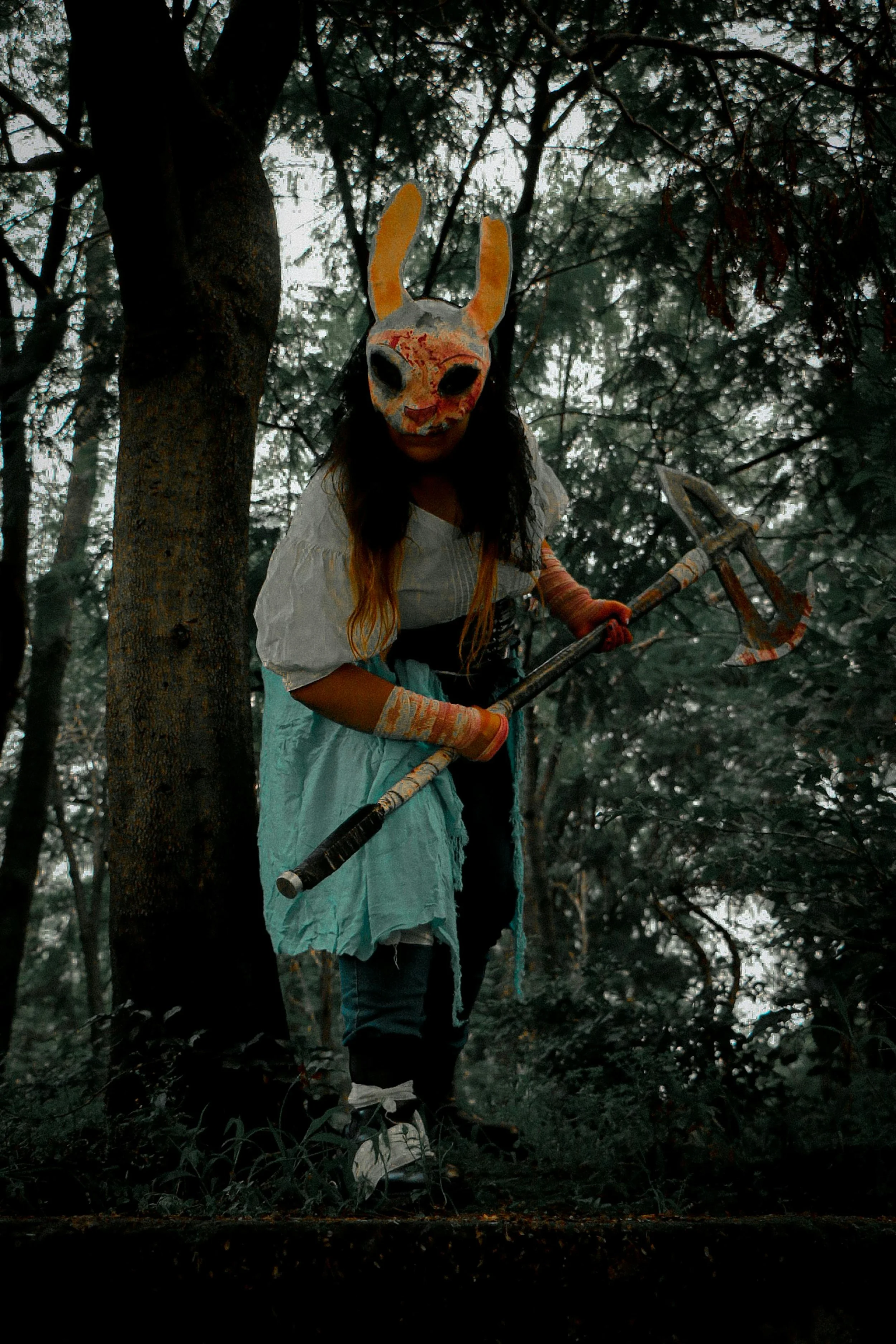 A person wearing a creepy rabbit mask with black eyes, a white shirt, and a blue skirt, holding a large axe, standing in a dark, misty forest surrounded by tall trees and dense foliage.