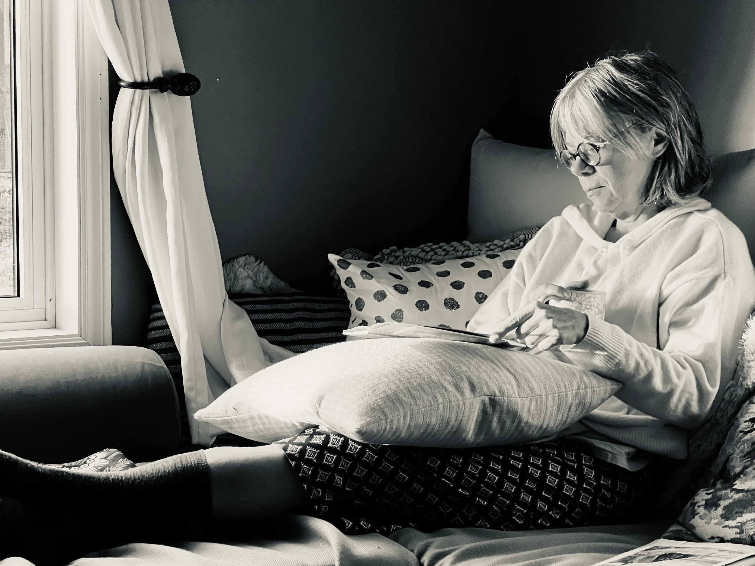 A woman with gray hair and glasses sitting on a couch, reading or looking at a device, with a window and curtain in the background.