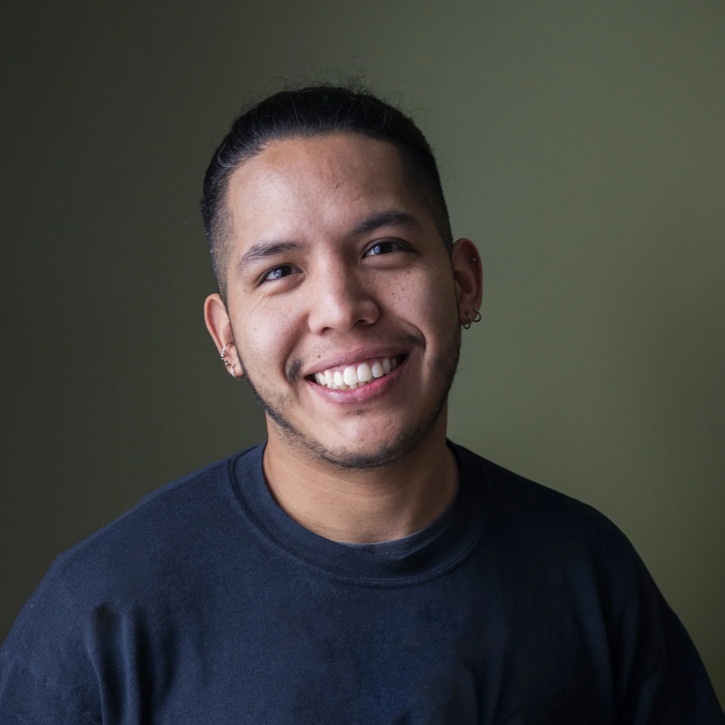 Close-up of a man smiling, wearing a navy blue shirt, with earrings, against a plain olive green background.