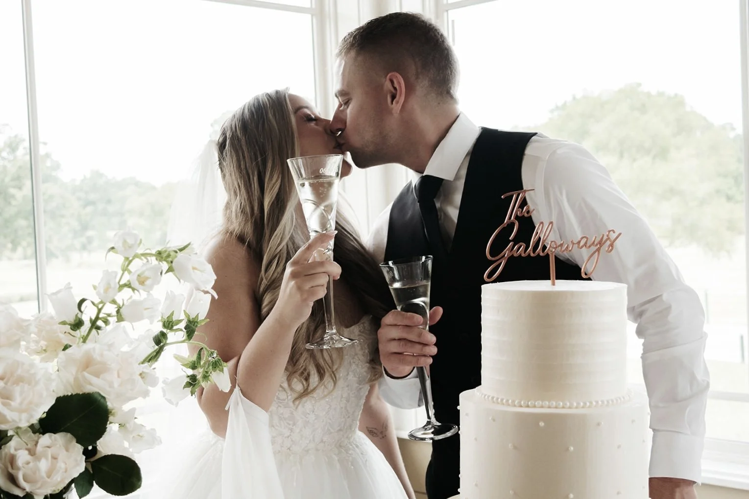 Bride and groom with champagne glasses and kissing by a 3 tier white wedding cake