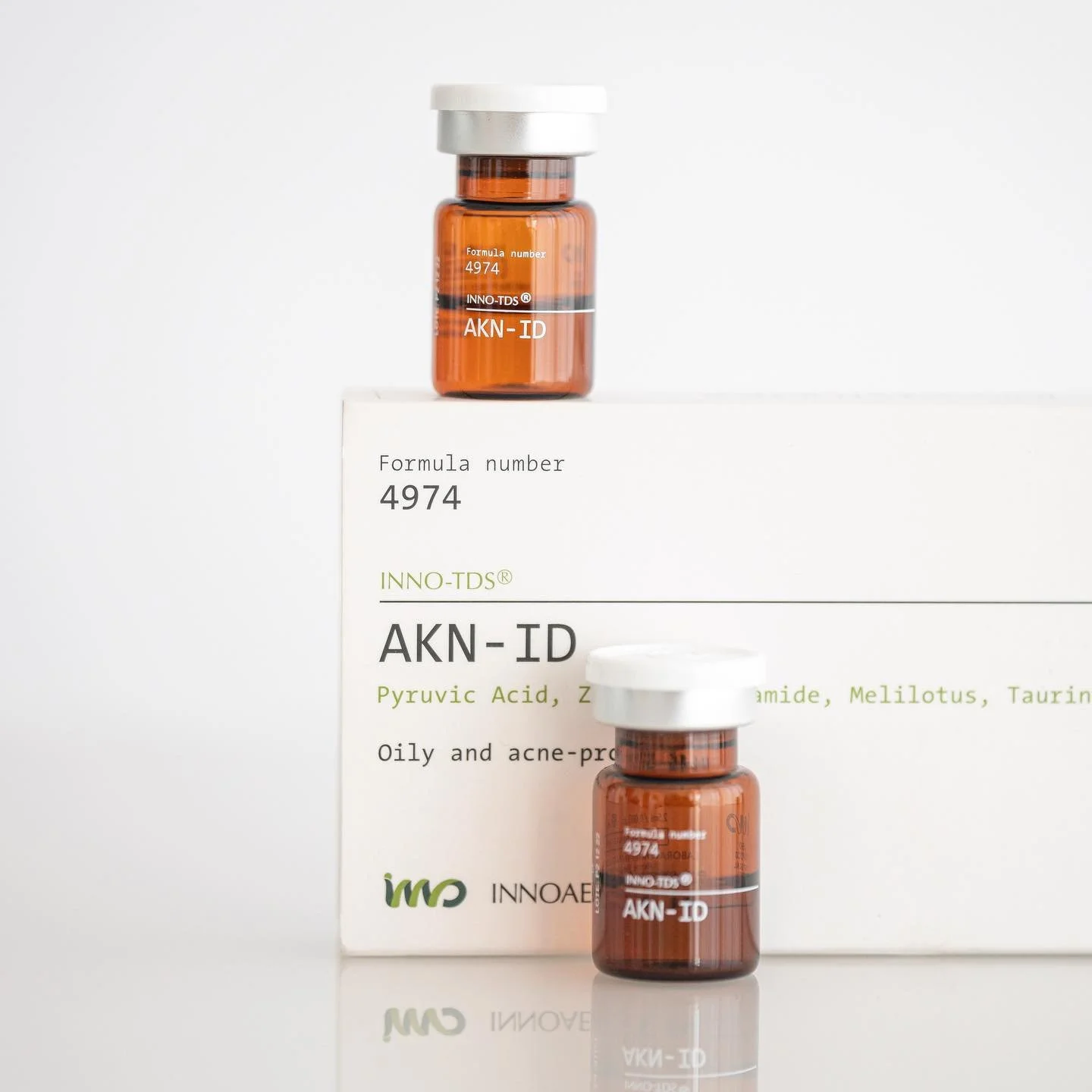 Two small amber glass bottles labeled 'AKN-ID' with white caps, placed on a white box with product information, against a plain white background.