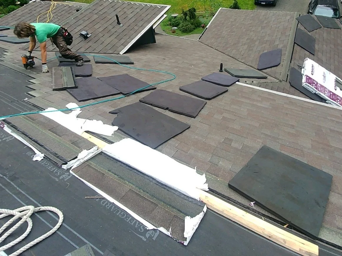 A person installing new shingles on a residential roof with several shingles already in place, surrounded by roofing tools and materials.